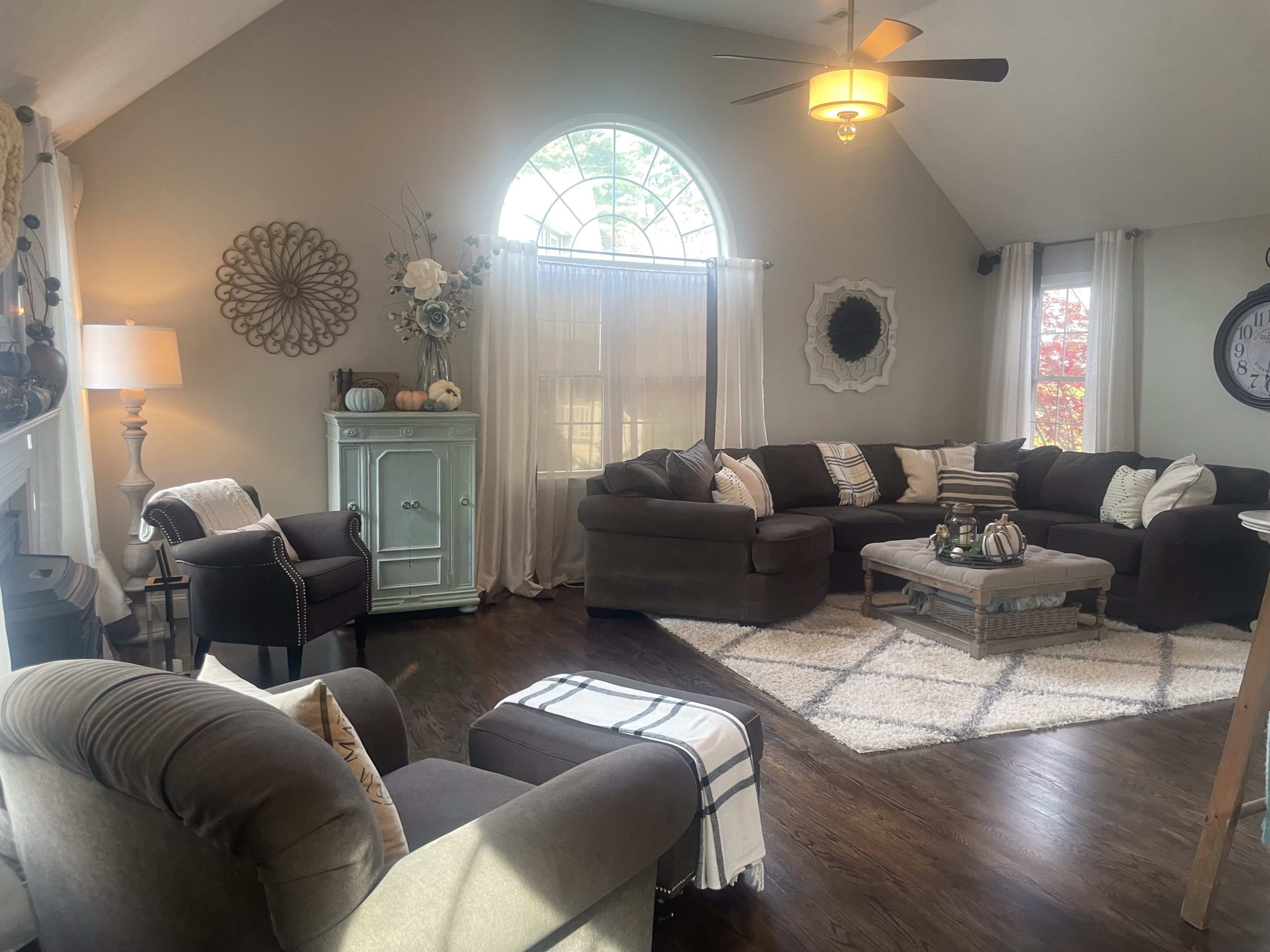 The living room features a large sectional sofa, a coffee table, and a window with sheer curtains, all arranged around a central area rug on hardwood flooring.