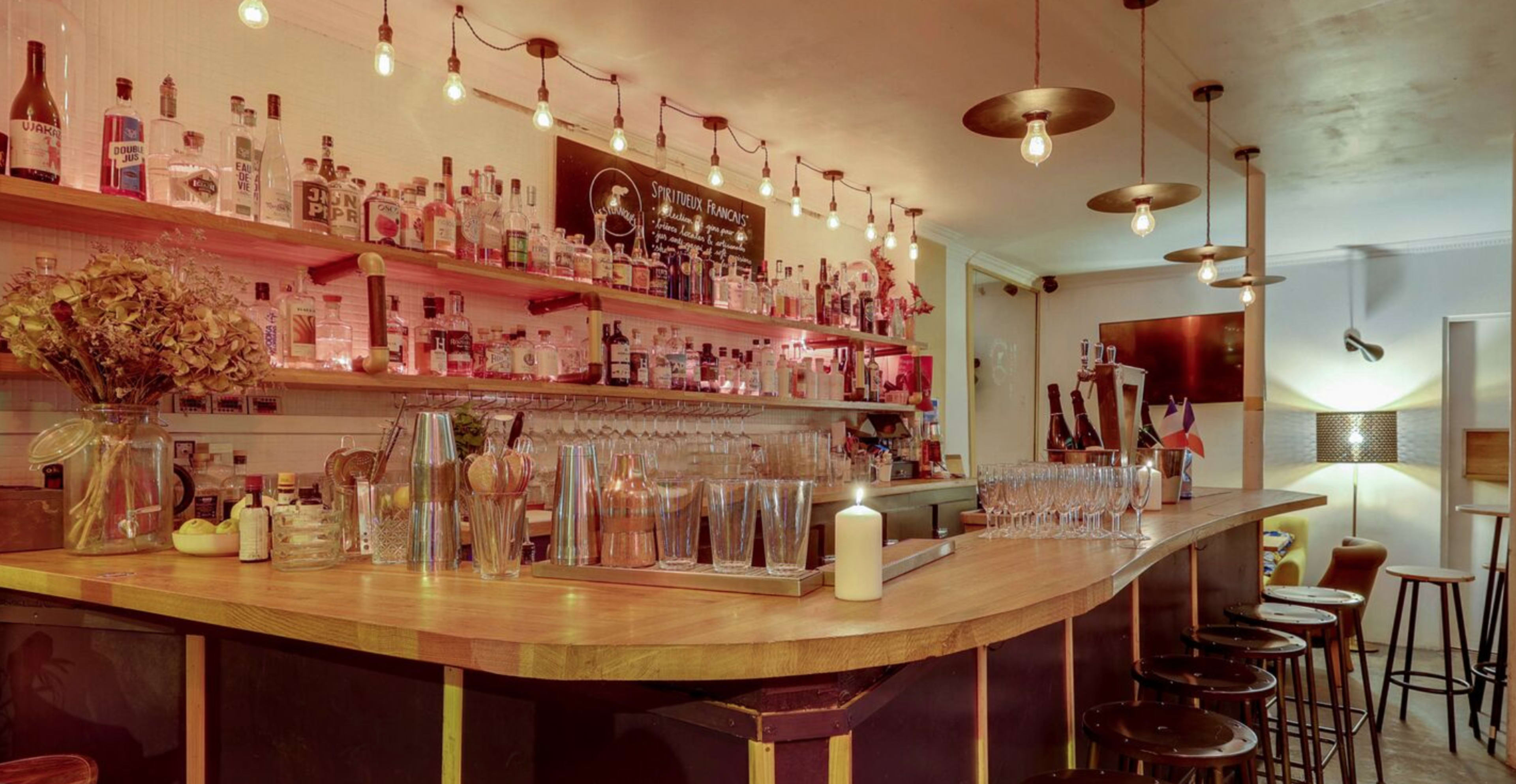 A well-lit bar with a wooden counter, shelves filled with various bottles, and high stools arranged in front of it.