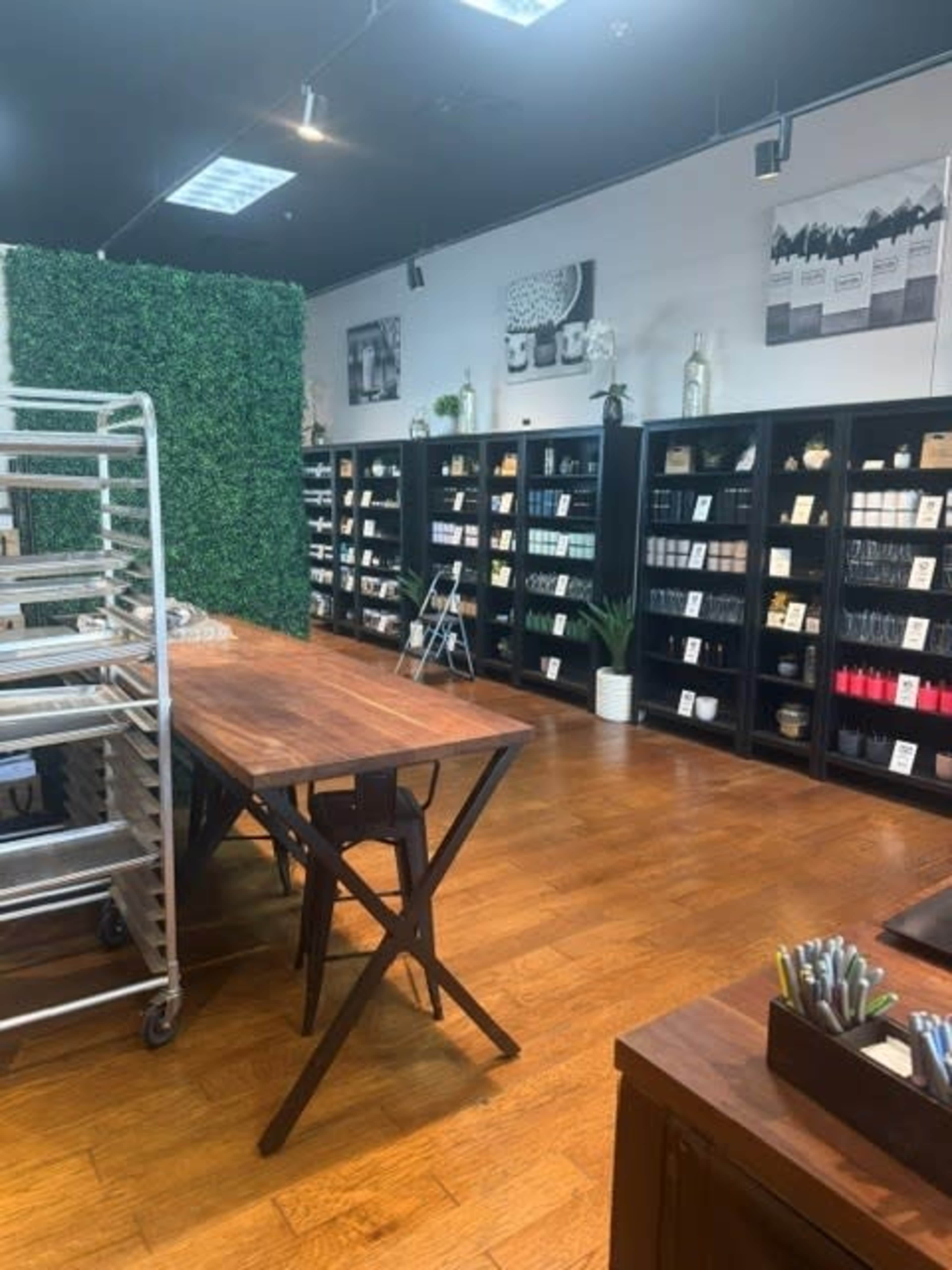 The image shows an interior of a store featuring wooden tables and shelves filled with various products, surrounded by greenery and minimal decorations.