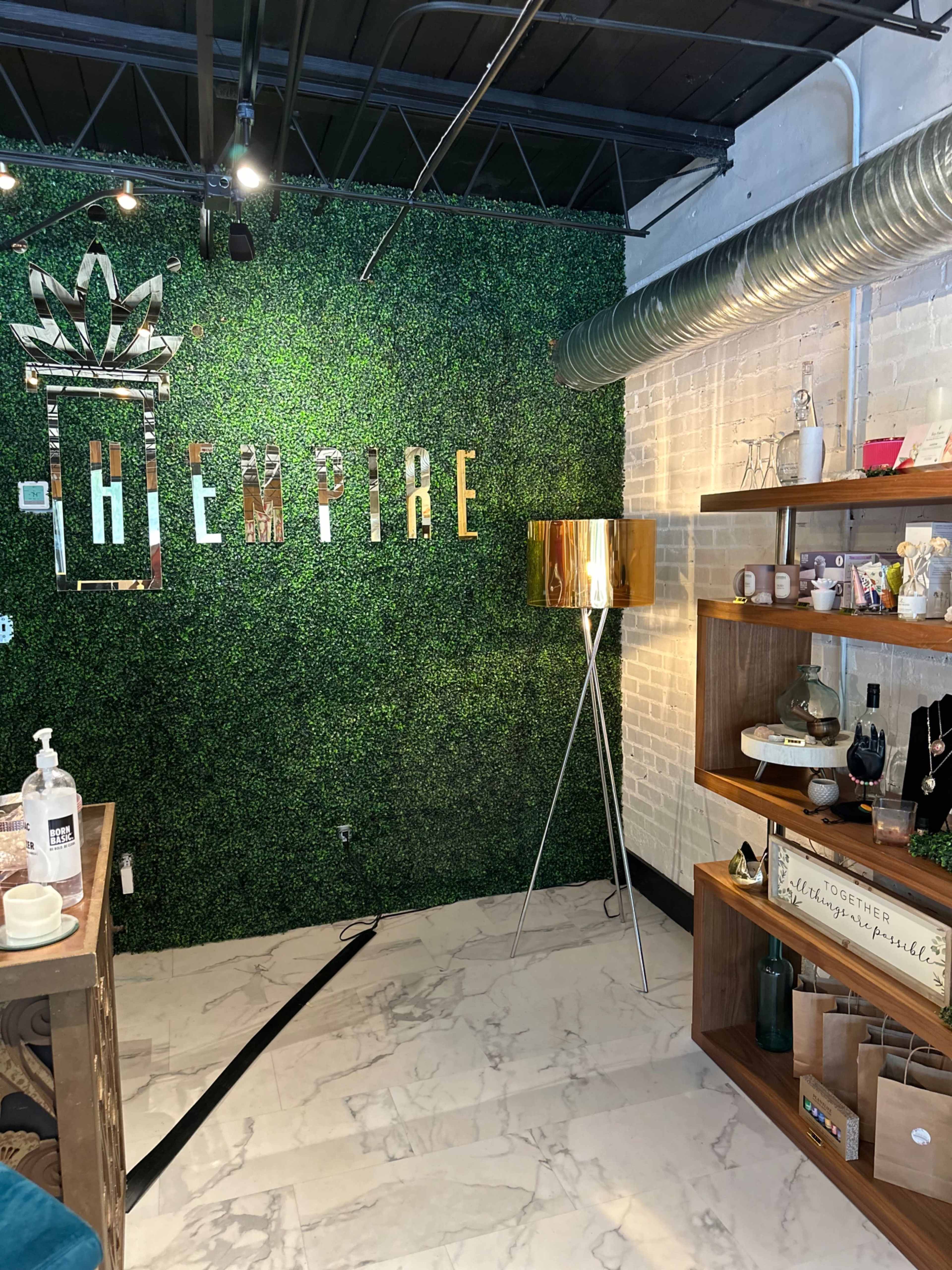 The image shows a retail space featuring a green, textured wall with the word "EMPIRE" in metallic lettering and a gold floor lamp beside wooden shelves filled with various decorative items.