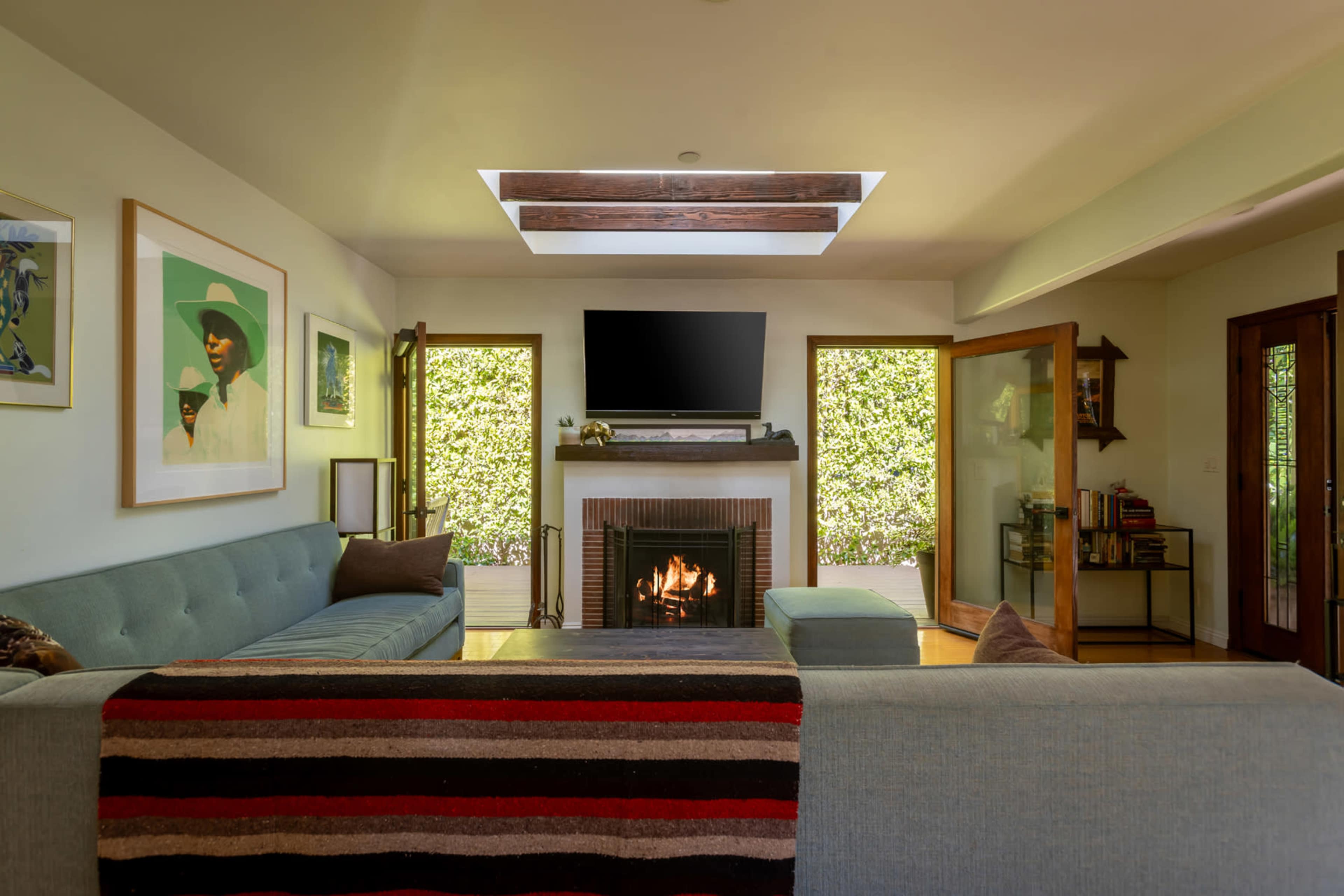 The image shows a modern living room with a fireplace, a large television mounted above it, and a sectional sofa, illuminated by natural light from a skylight and windows.
