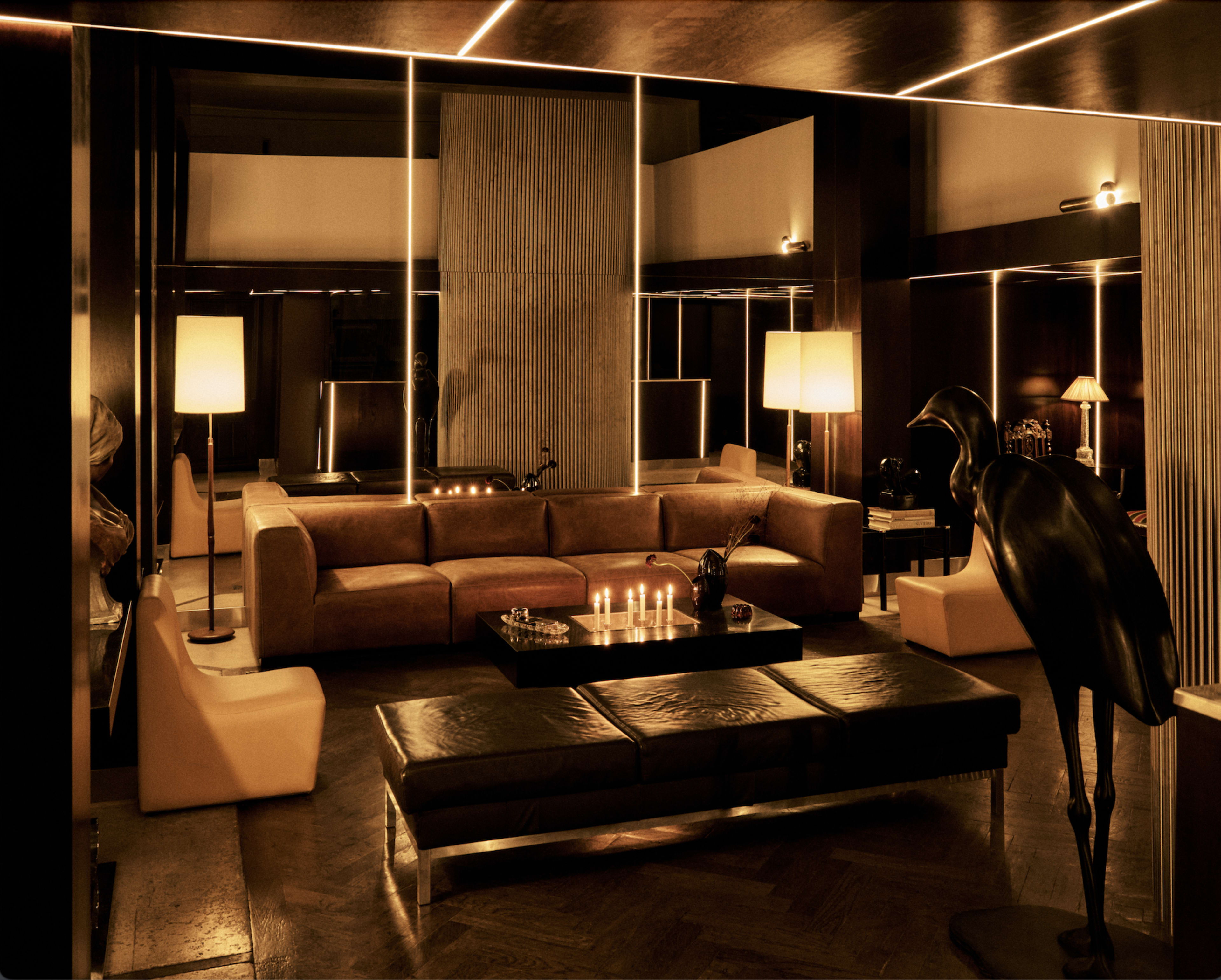 The image depicts a modern, dimly lit lounge area featuring a large brown sofa, a black coffee table with candles, and several lamps illuminating the space.