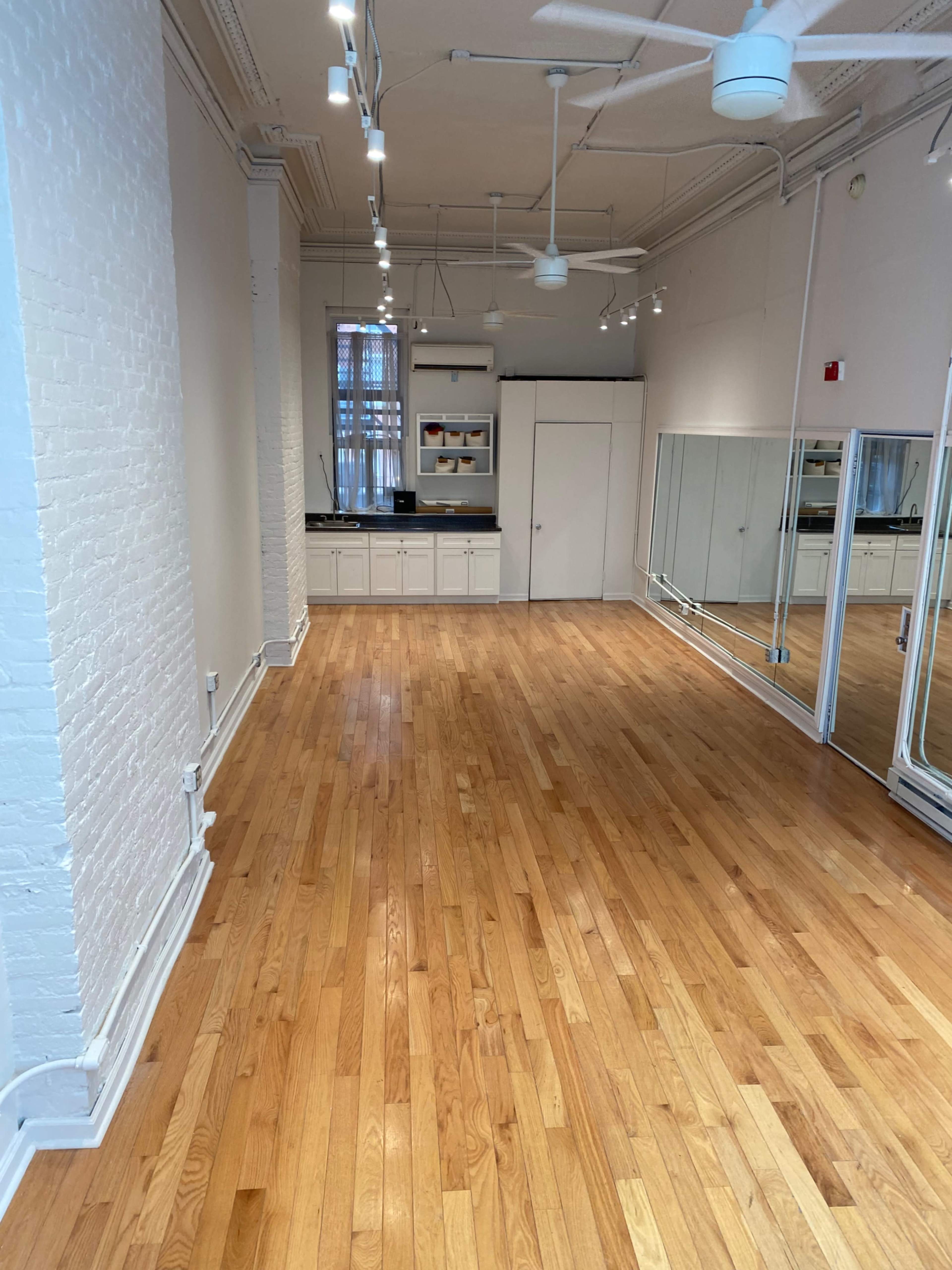 An empty room with polished wooden flooring, white brick walls, and a mirrored wall at one end, along with a kitchenette area featuring cabinets.