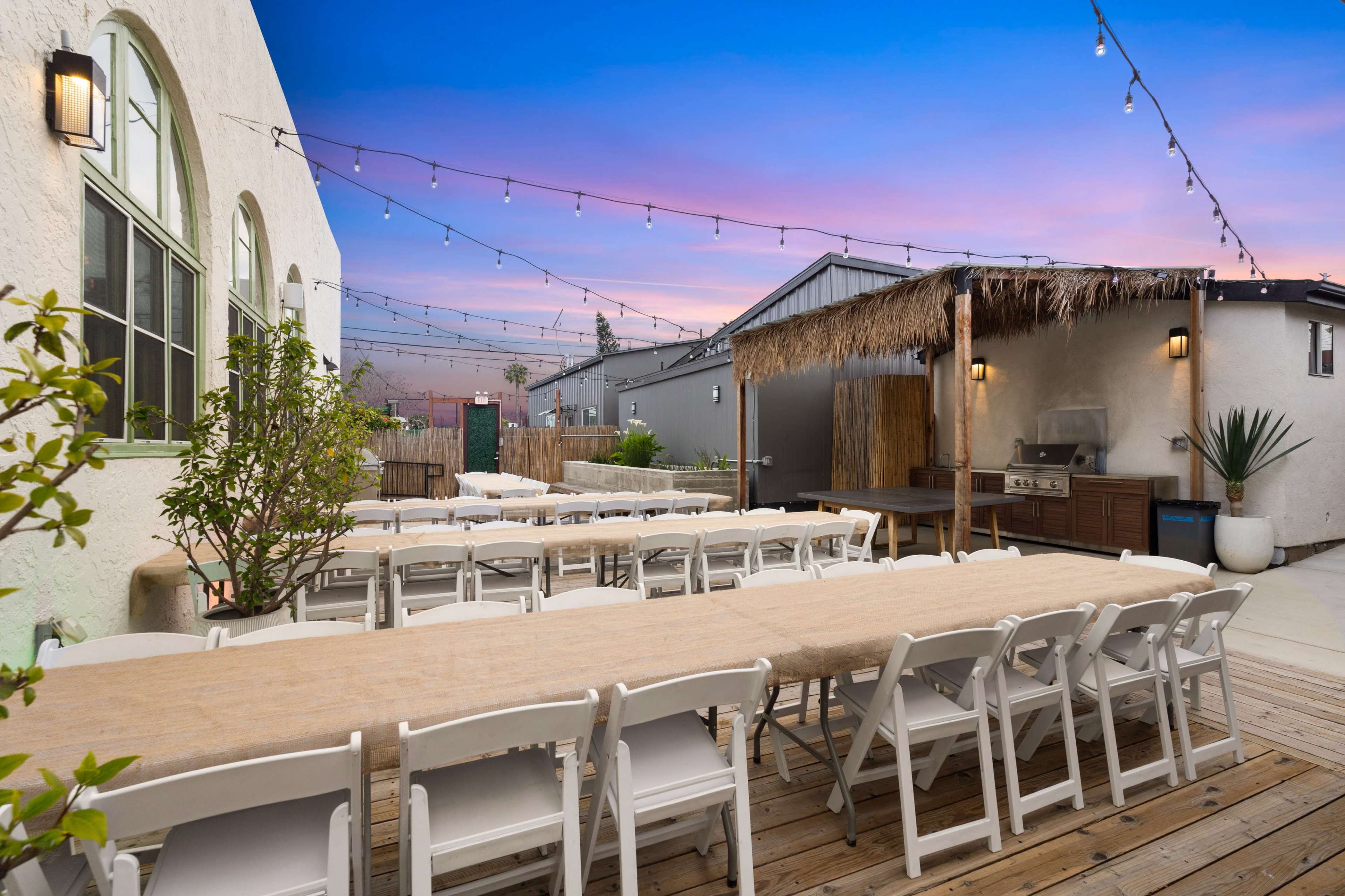 A well-arranged outdoor seating area with long tables set up under string lights, adjacent to a barbecue area and surrounded by a landscaped space at dusk.