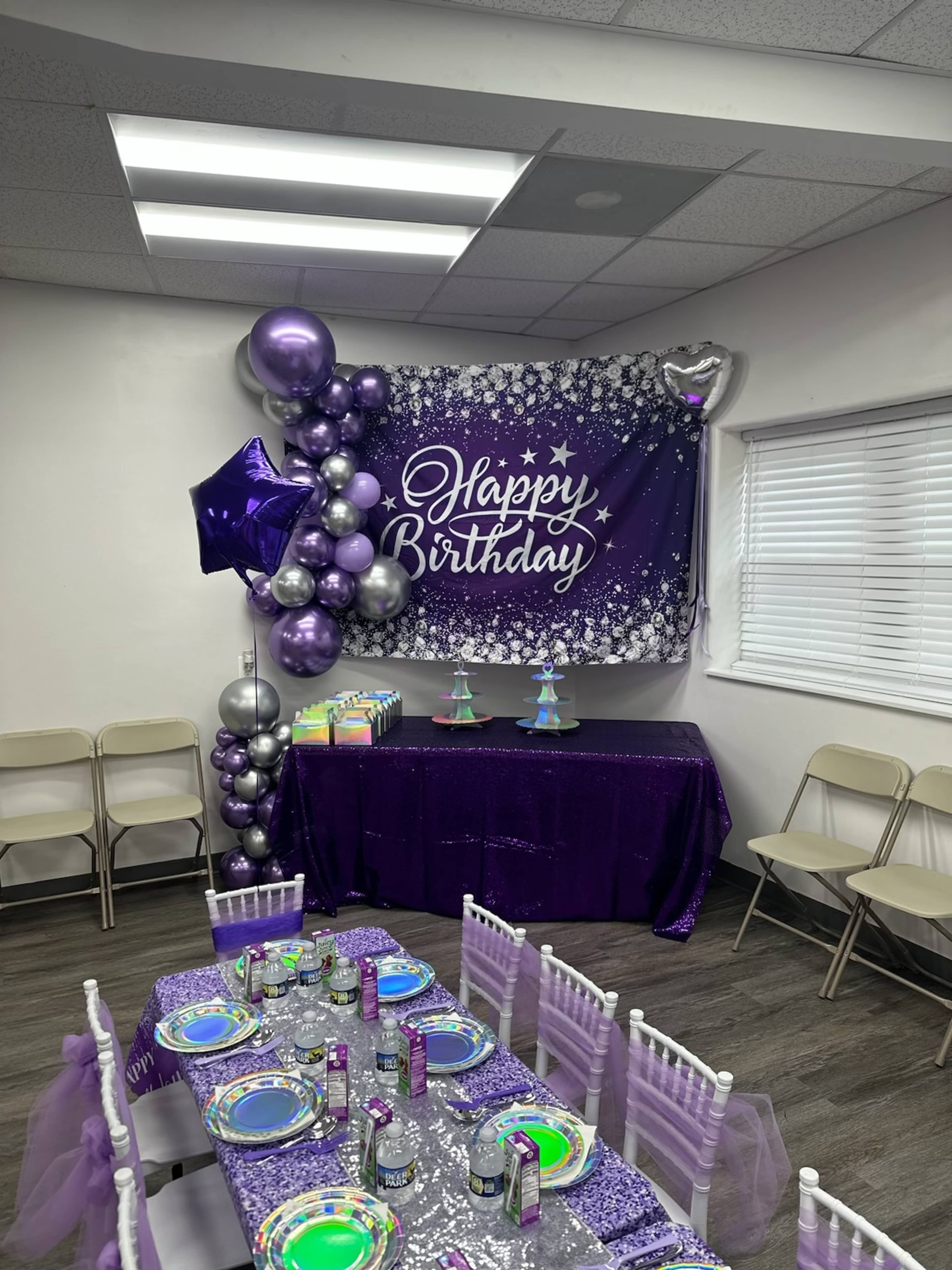A birthday celebration setup featuring a "Happy Birthday" banner, purple decorations, a table with colorful plates, and a dessert display.