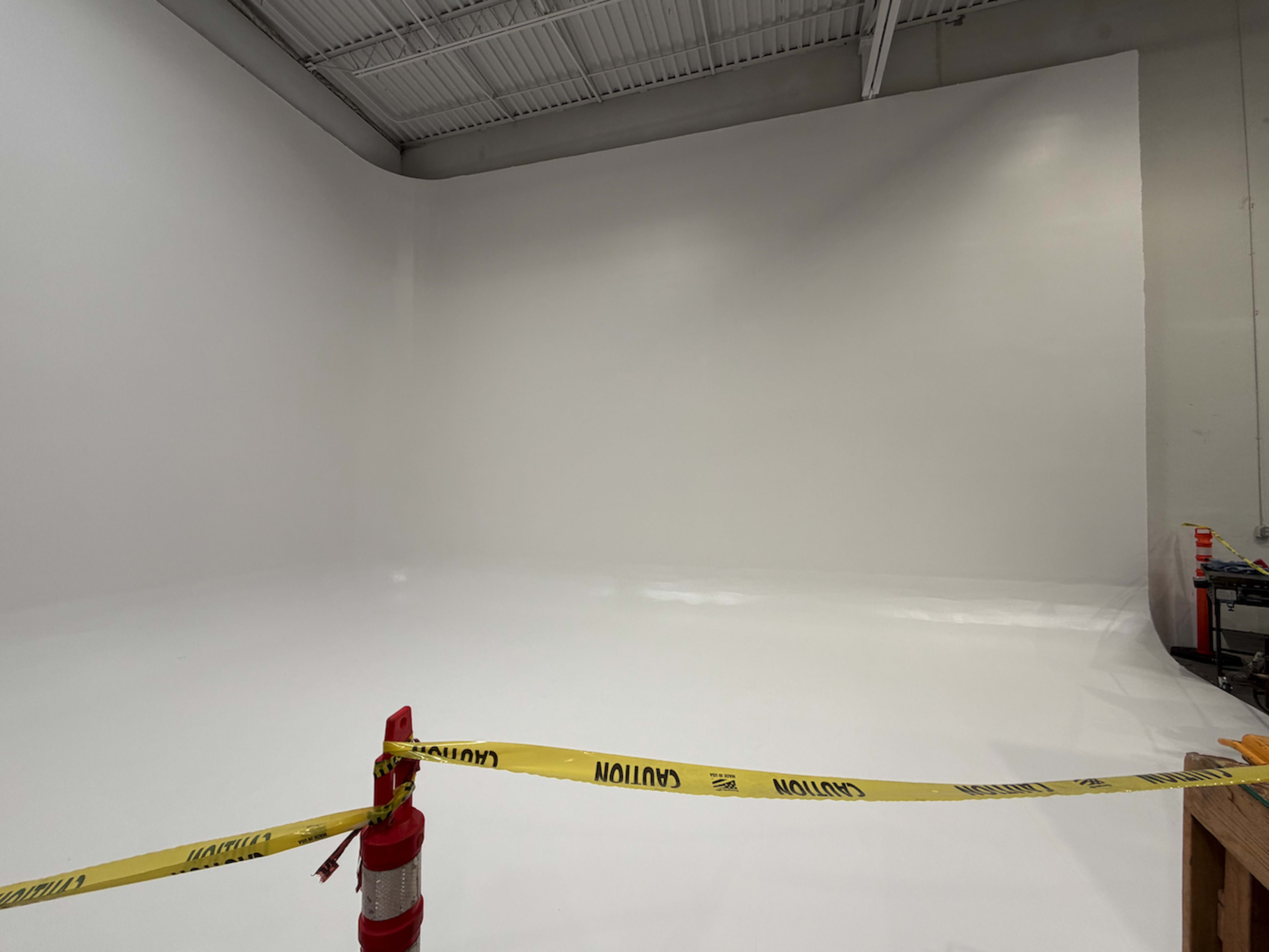 The image shows a large, empty studio space with white walls and a shiny floor, surrounded by caution tape and a traffic cone.