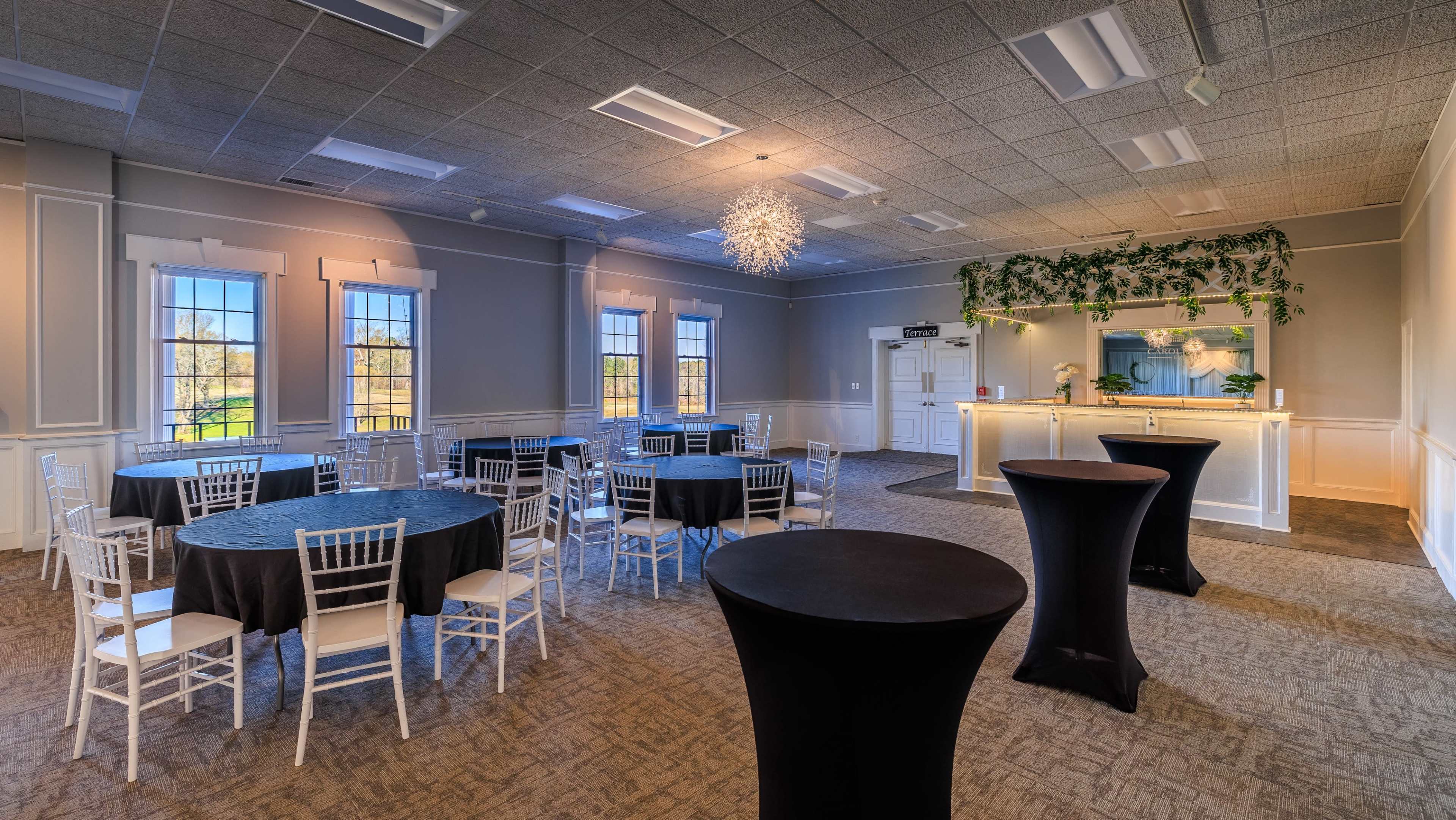 A spacious event room with round and tall tables, elegant white chairs, and a counter adorned with greenery and lighting.