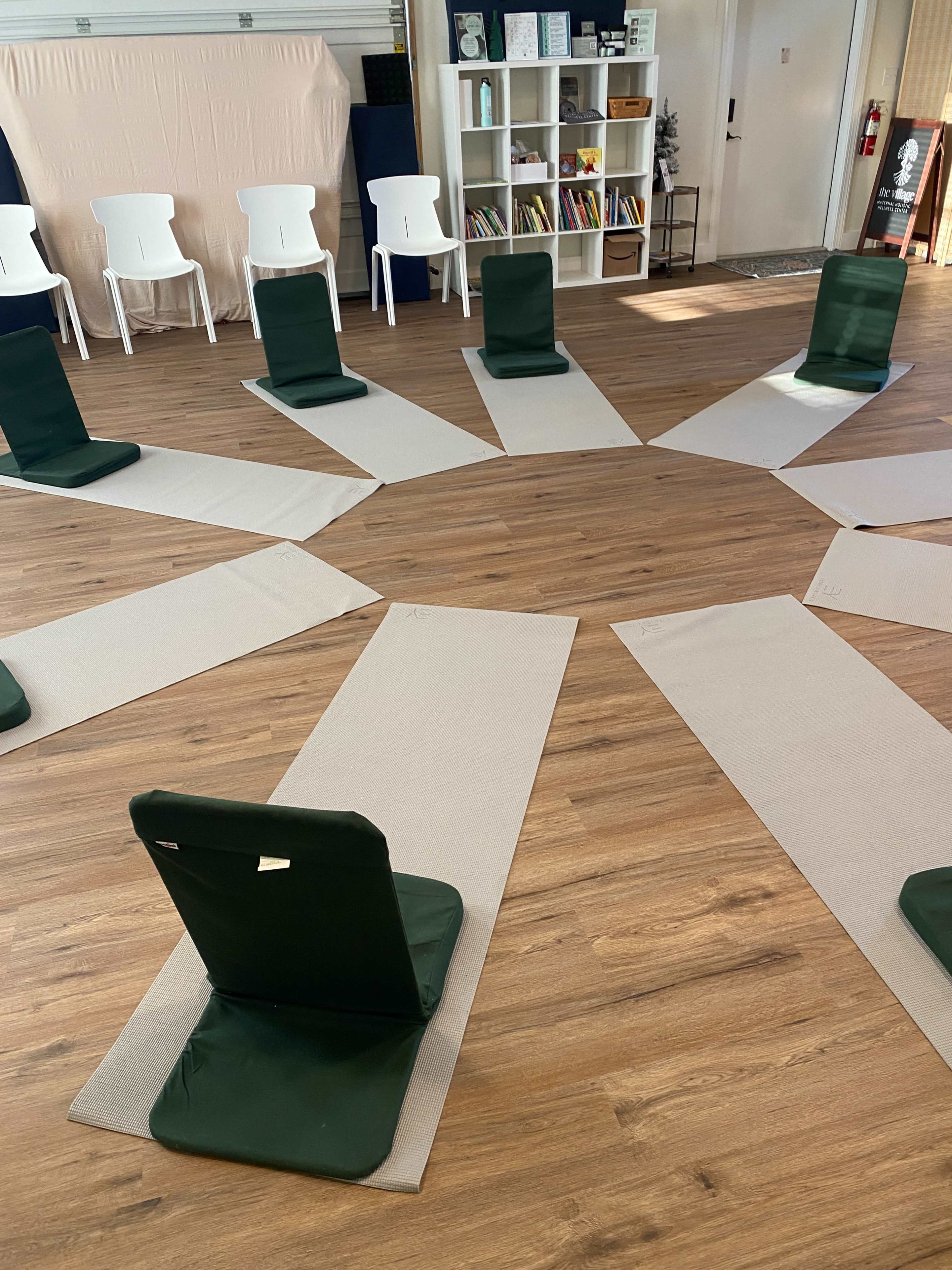 The image shows a circular arrangement of green chairs on gray mats in a spacious room with light wooden flooring and shelves in the background.
