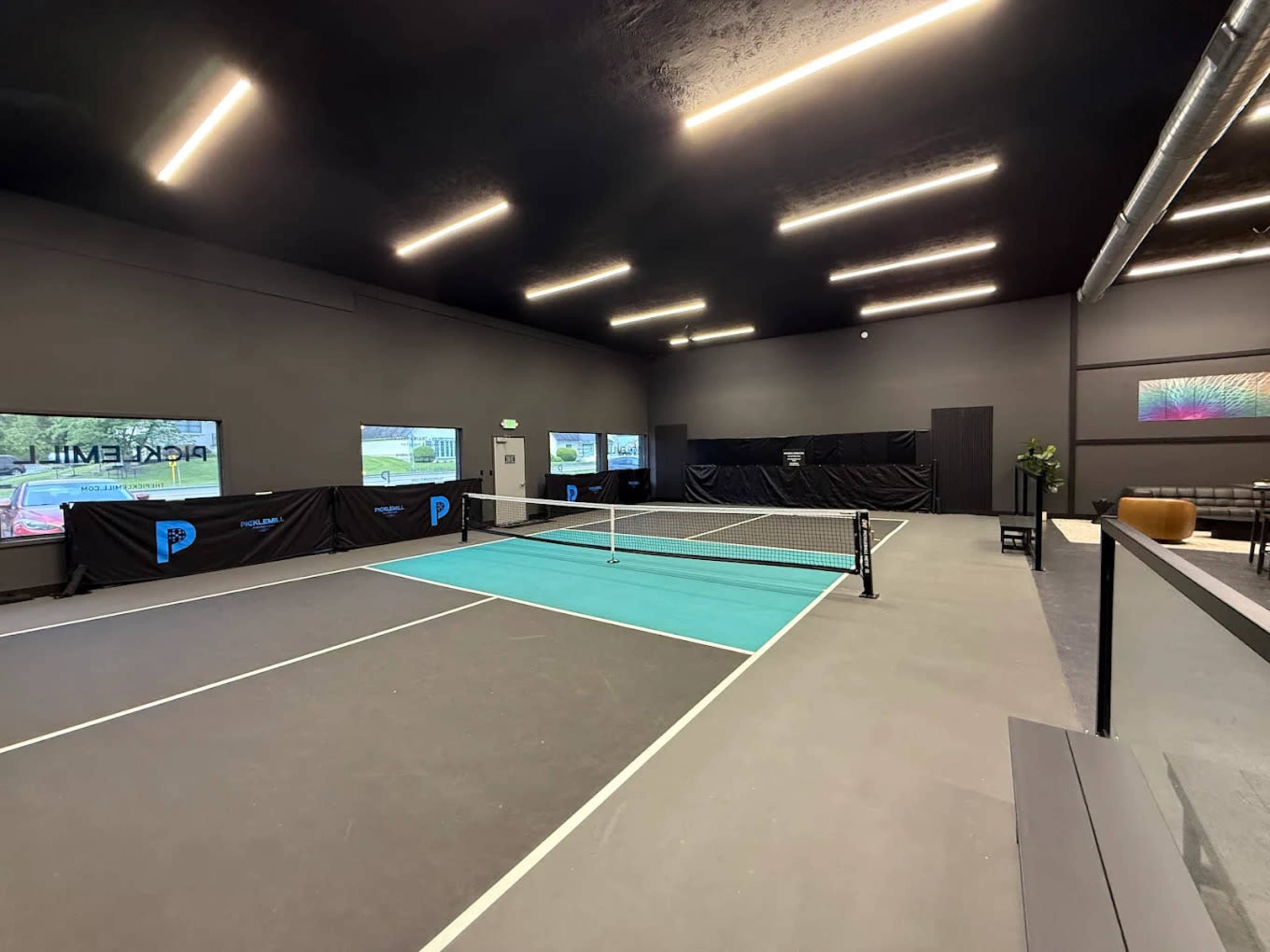 The image shows a modern indoor tennis court, featuring a teal center line, surrounded by black walls and digital screens displaying images.