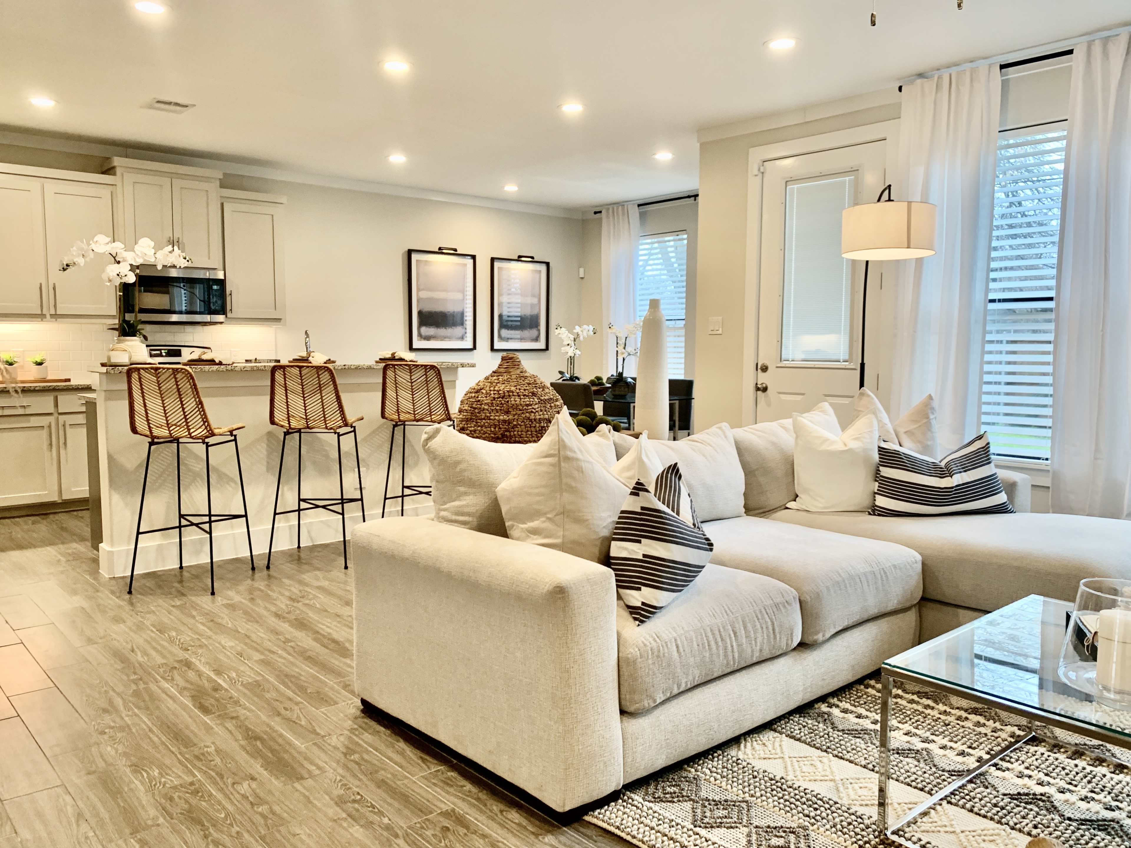 A modern living space with a light-colored sectional sofa, a kitchen area featuring bar stools, and large windows with sheer white curtains.