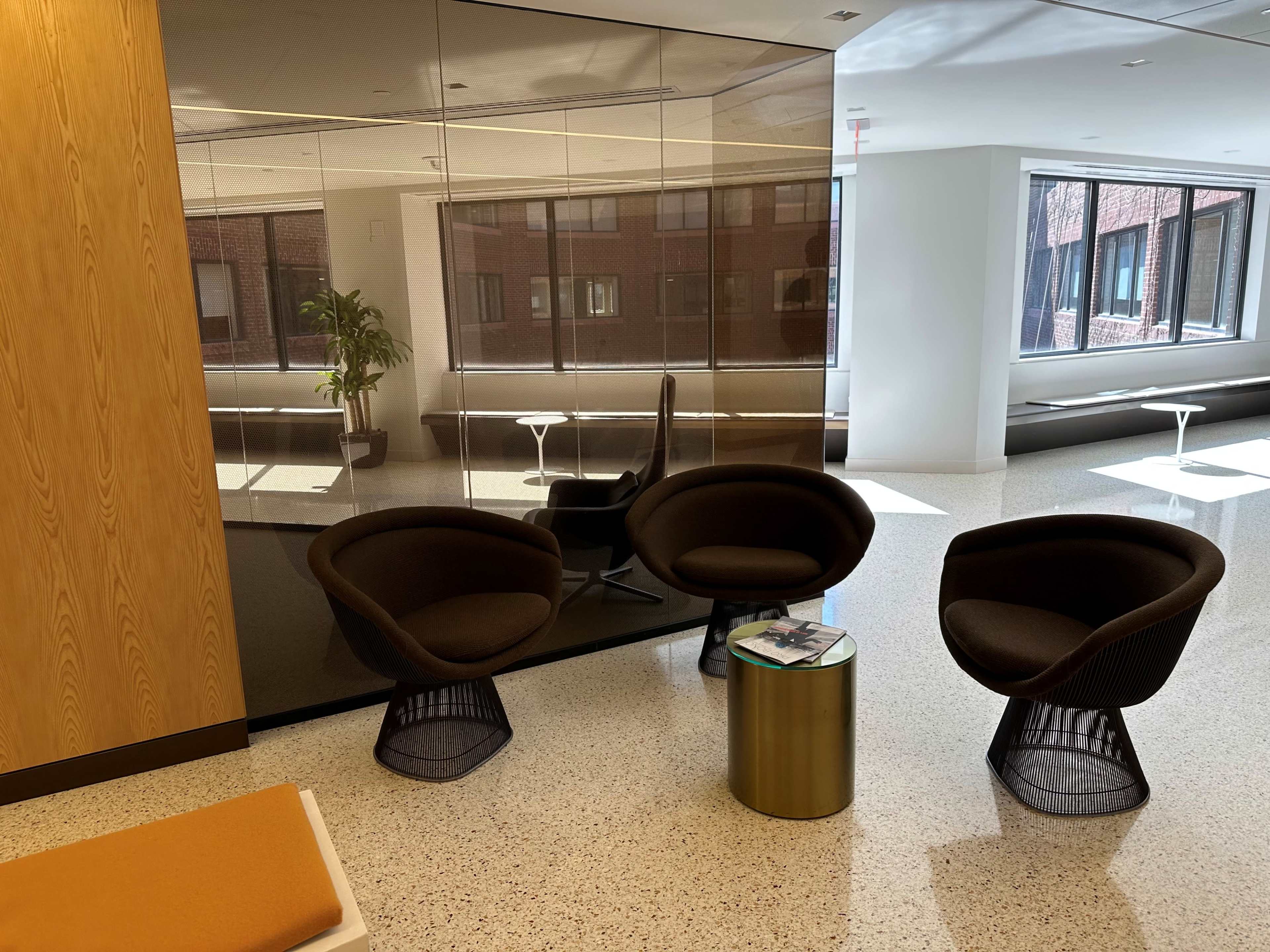 The image shows a modern seating area featuring three curved chairs around a small round table, with large windows and a reflective wall in the background.