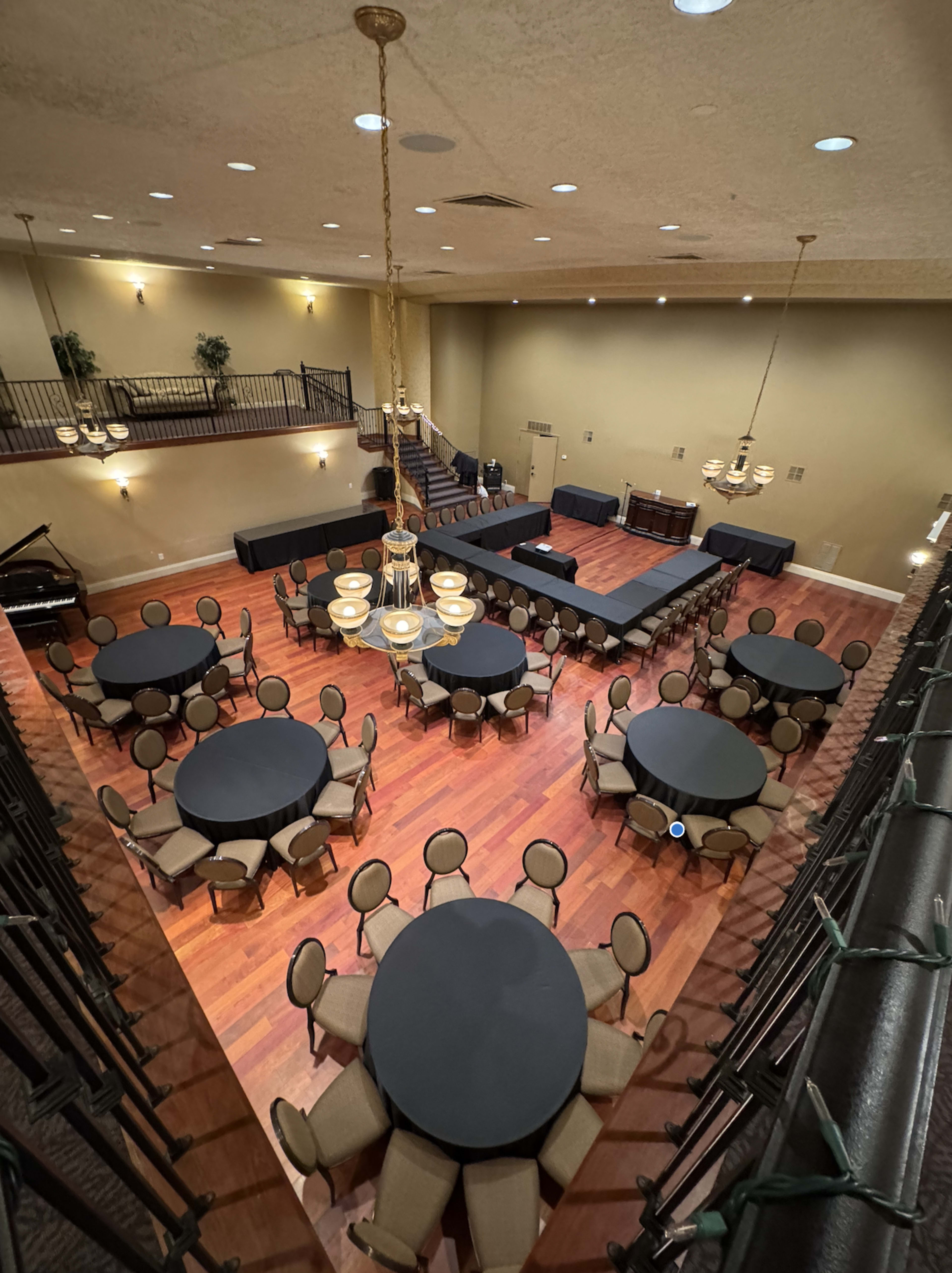 The image shows a spacious event room with multiple round tables set up, arranged around a central stage area, and features a grand chandelier.