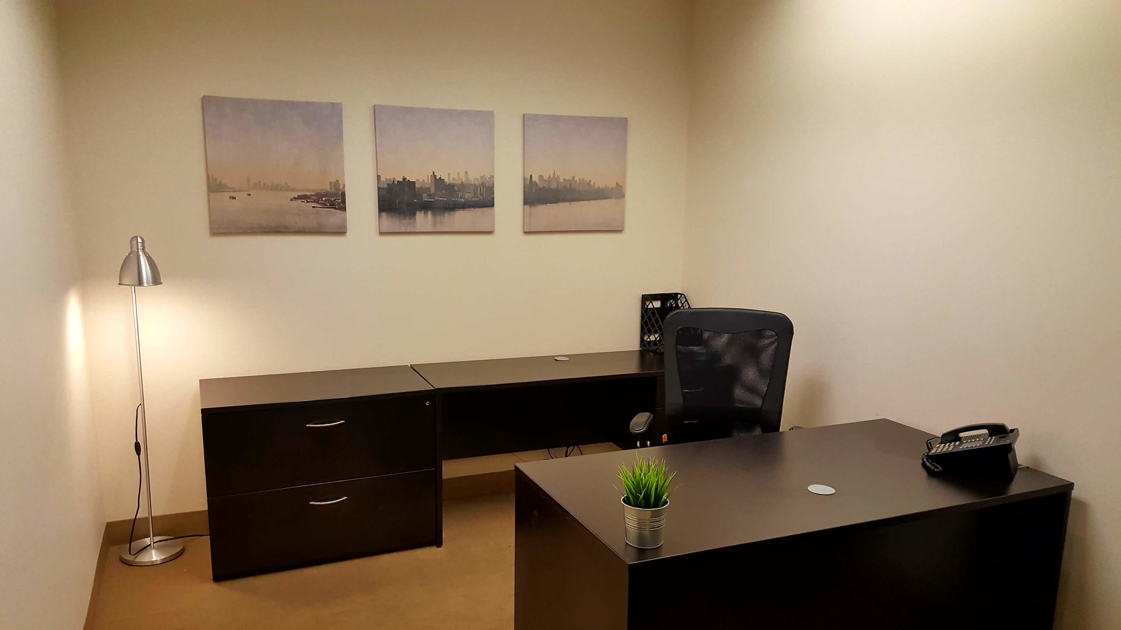 A minimalistic office space featuring two black desks, a chair, a lamp, a telephone, and three framed cityscape prints on the wall.
