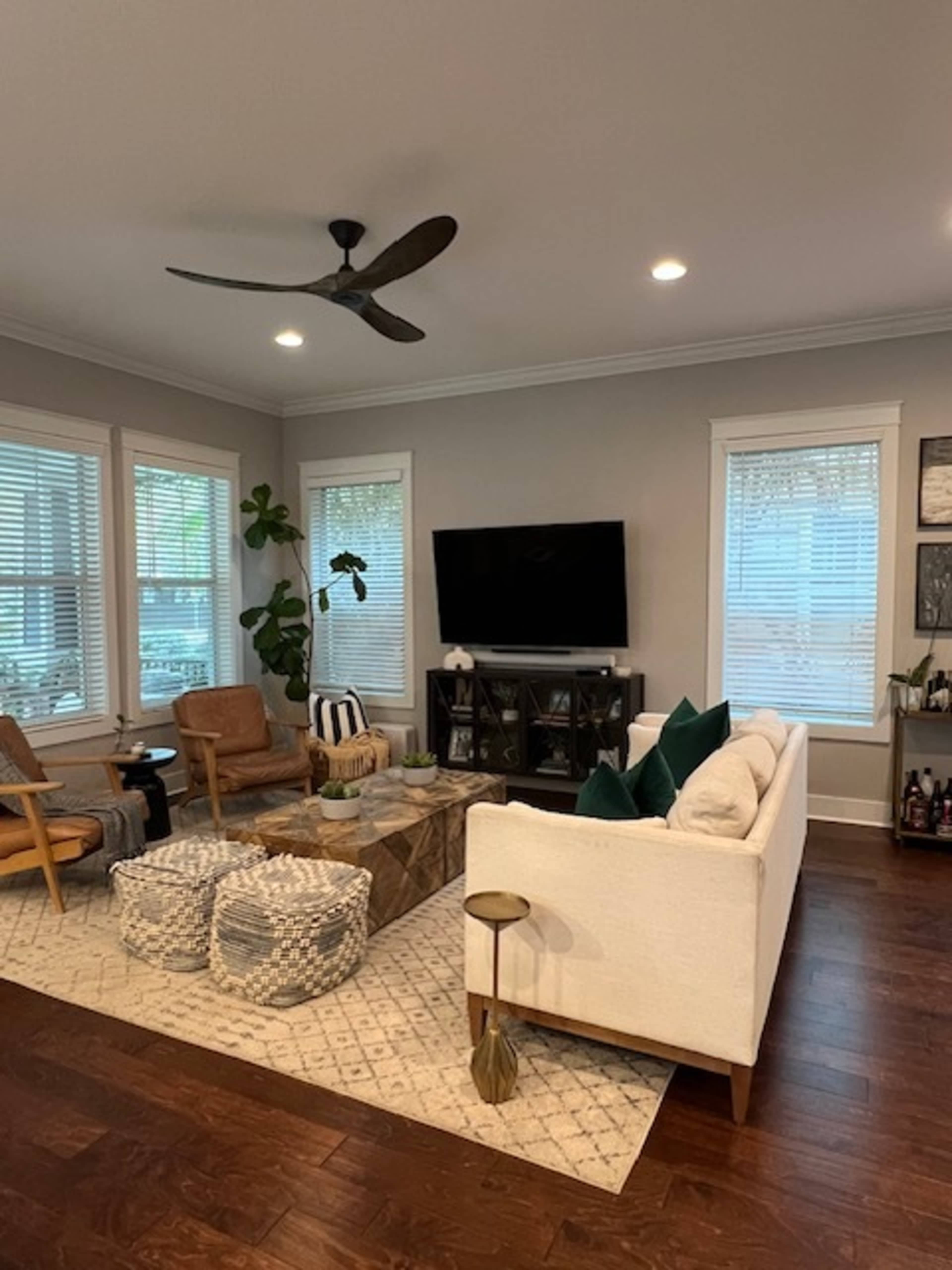 A neatly arranged living room features a white couch, wooden coffee table, and various seating options, with a television mounted on the wall and windows allowing natural light.