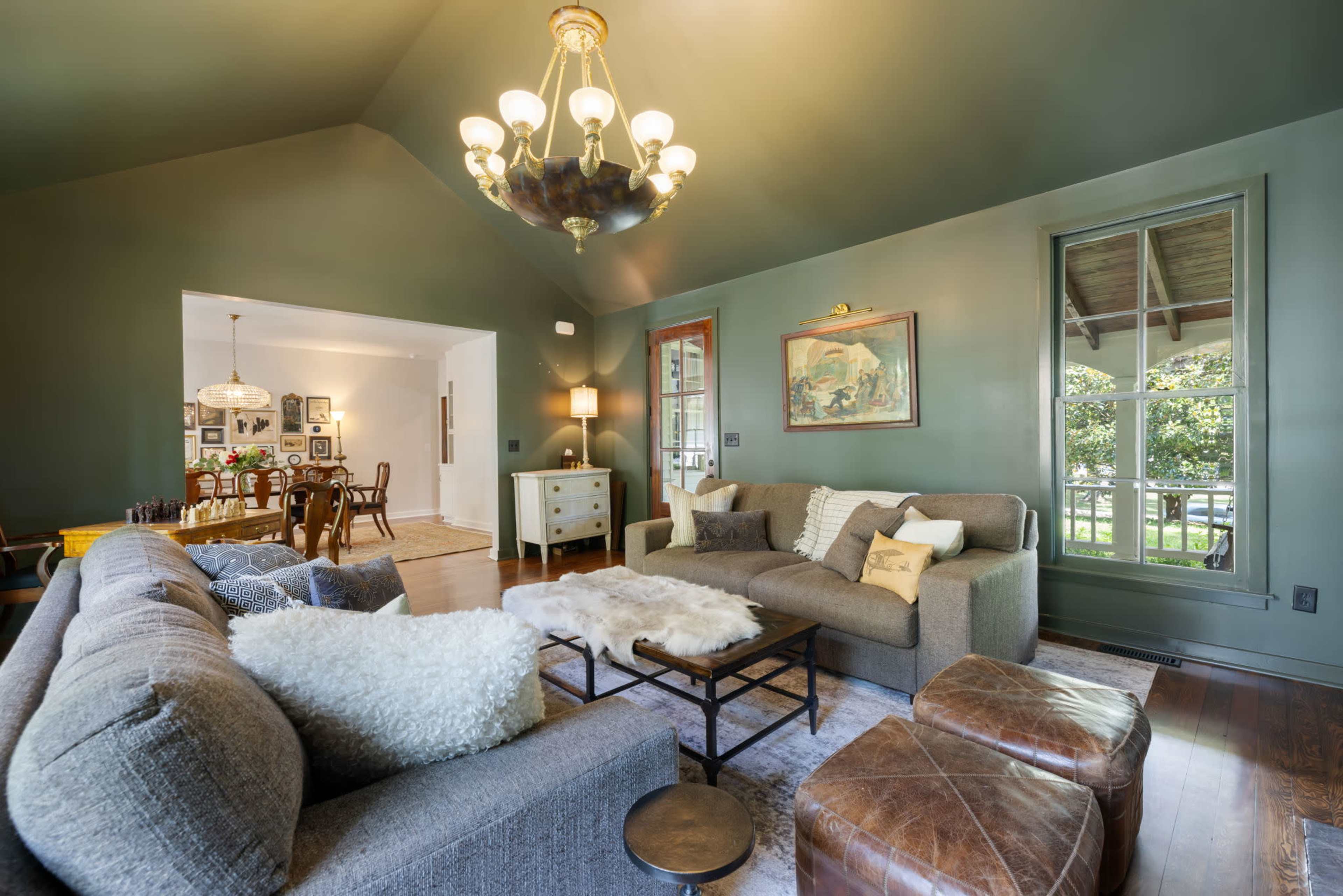 The image shows a living room with green walls, a chandelier, and a mix of seating, including a gray sofa and leather ottomans.