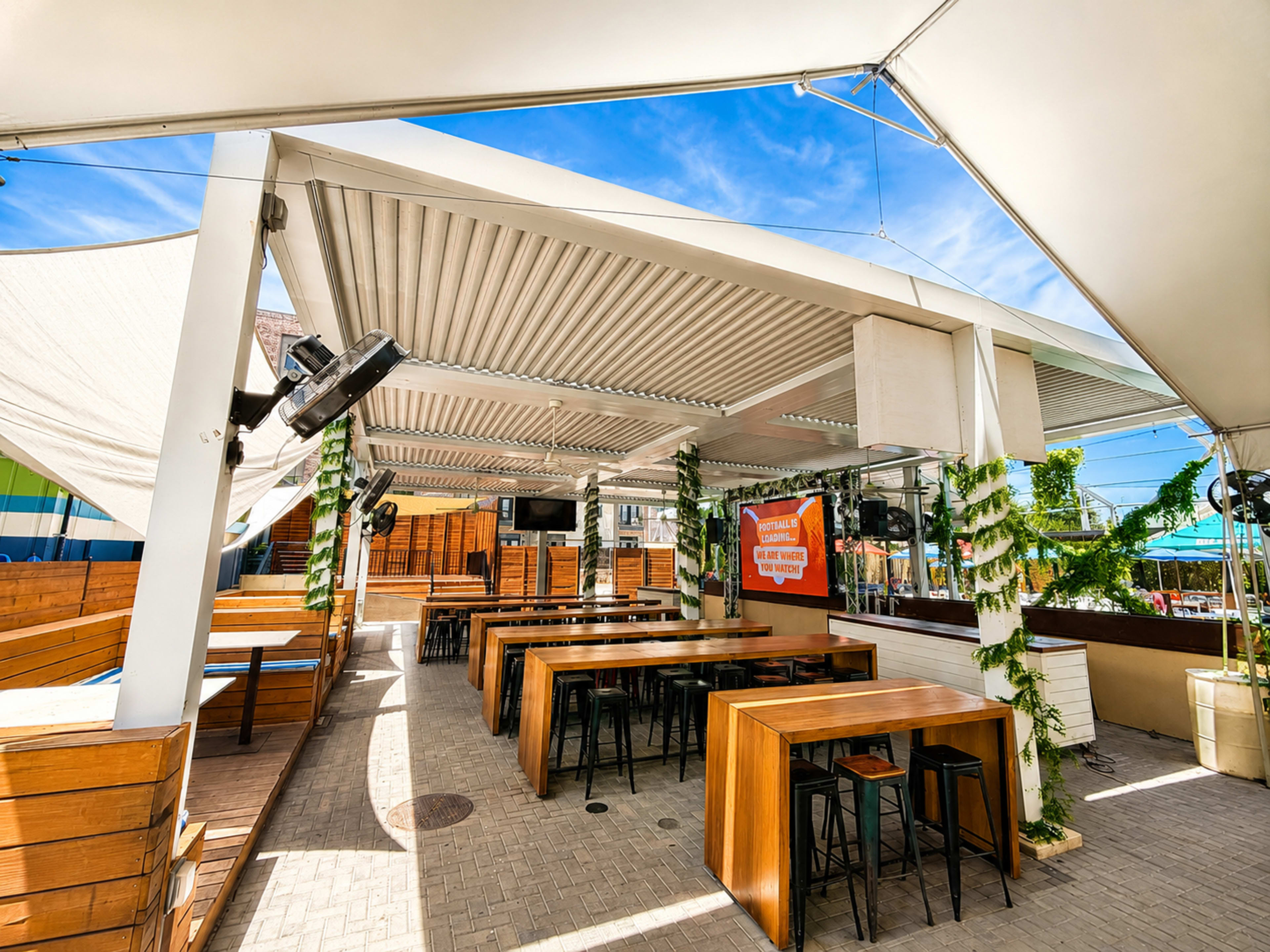 The image shows a spacious outdoor dining area with wooden tables and benches, covered by a large canopy, and featuring various lights and a promotional sign.