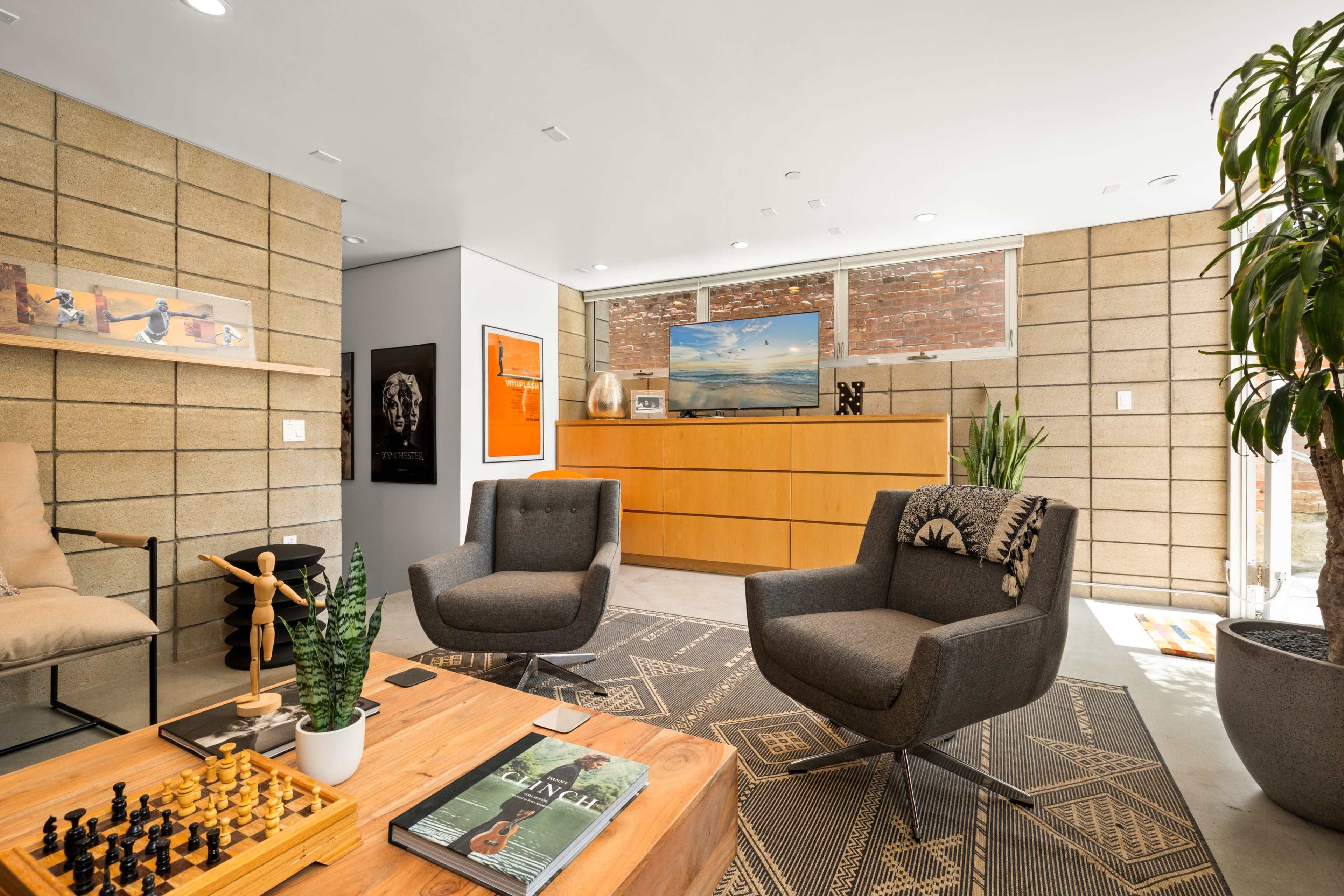 The image shows a modern living room with two gray chairs, a wooden coffee table featuring a chess set, and a large wooden cabinet against a wall of exposed bricks and windows.