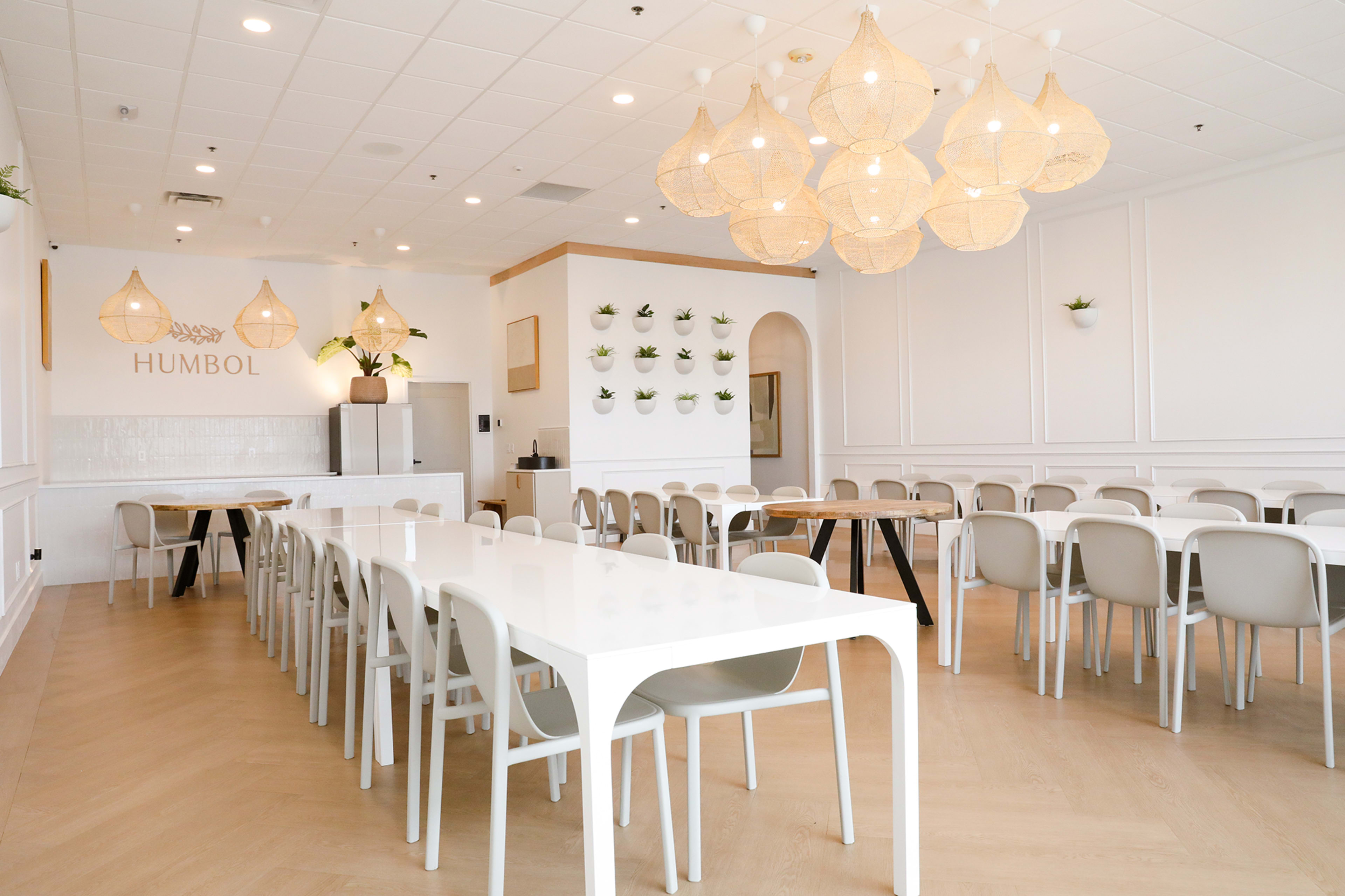 A modern, well-lit dining area with white tables and chairs, featuring decorative pendant lights and greenery on the walls.