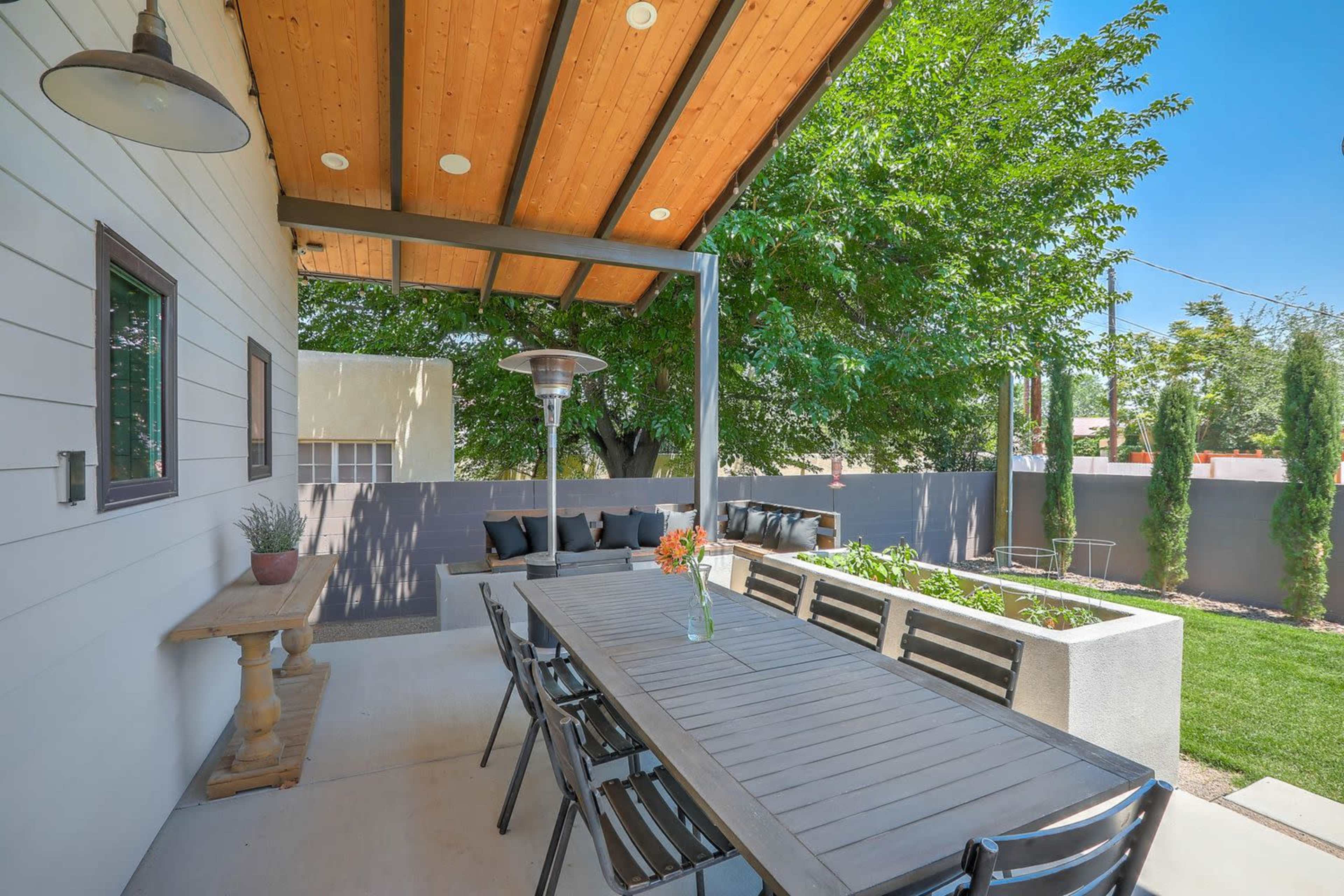 A patio area features a long dining table under a wooden roof, surrounded by greenery and seating.