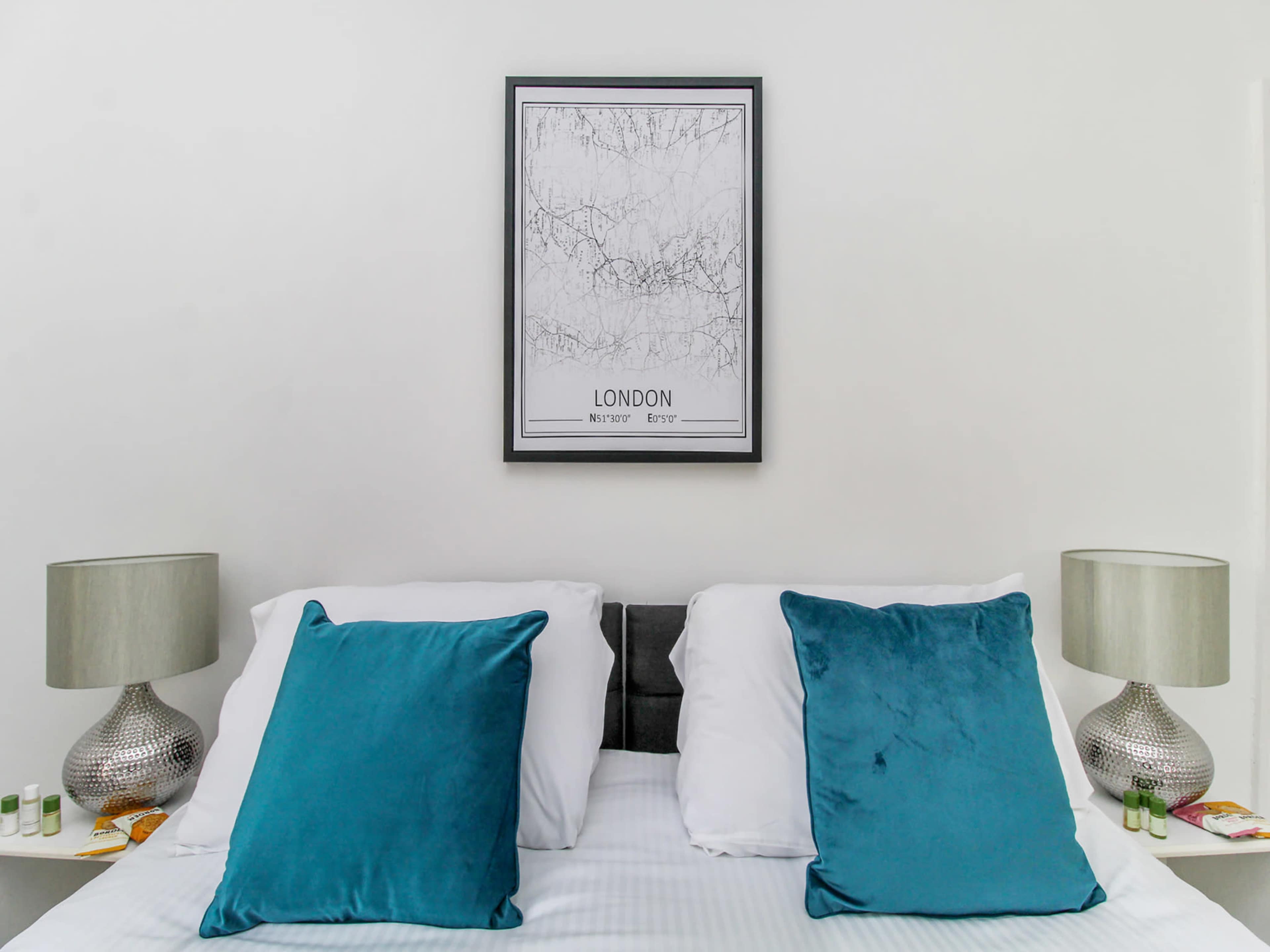 The image shows a neatly arranged bed with white linens and two teal pillows, flanked by silver table lamps and a framed map of London on the wall above.