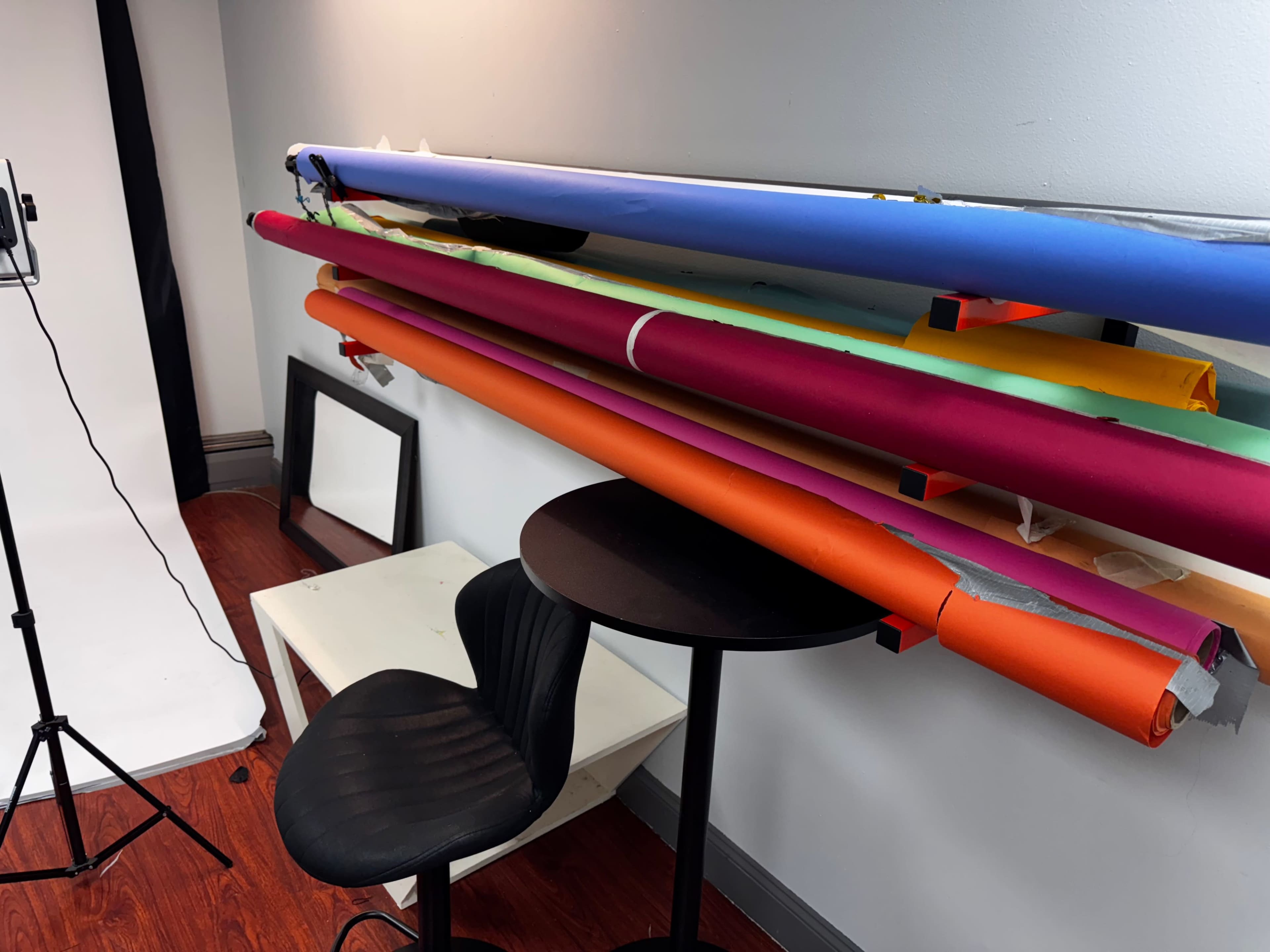 The image shows a studio space with colorful backdrop rolls hanging on a wall above a black chair and a small table.