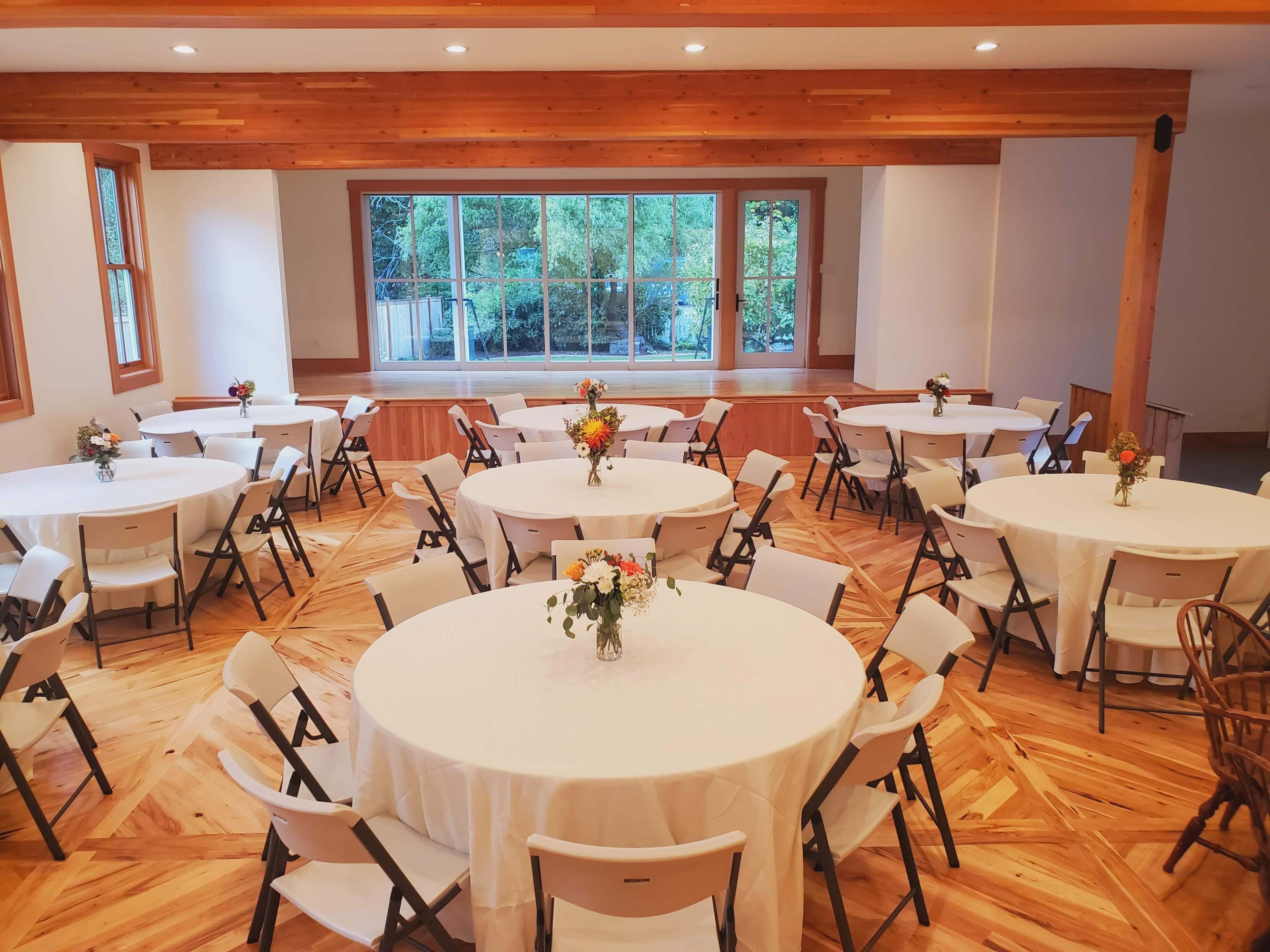 A spacious indoor event room with several round tables set with white tablecloths and floral centerpieces.