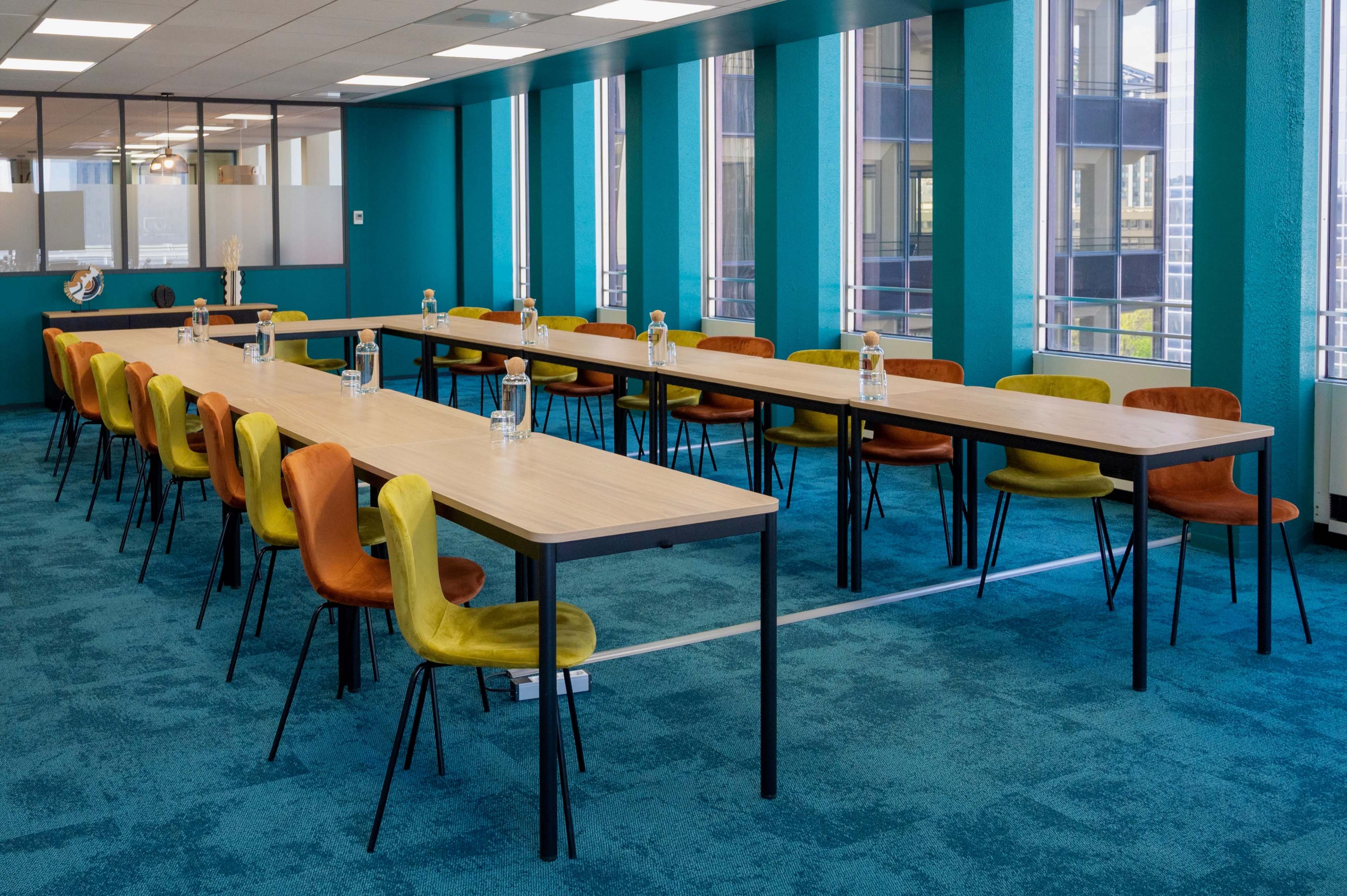 A spacious meeting room features long tables arranged in a row, each accompanied by colorful chairs and bottles of water.