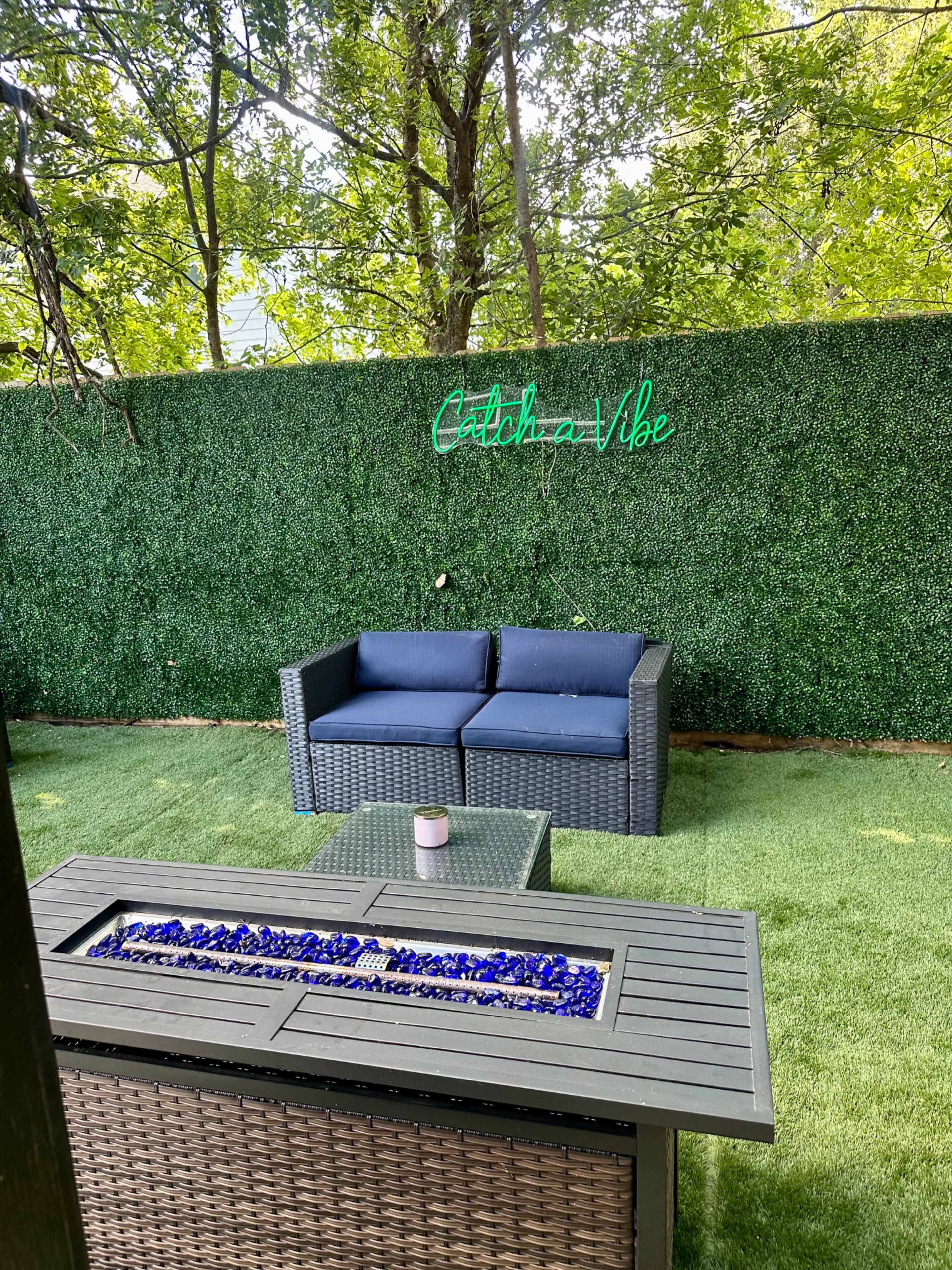 A cozy outdoor space features a rattan couch with navy cushions beside a fire pit adorned with blue glass stones, under a green hedge and a neon sign that reads "Catch a Vibe."