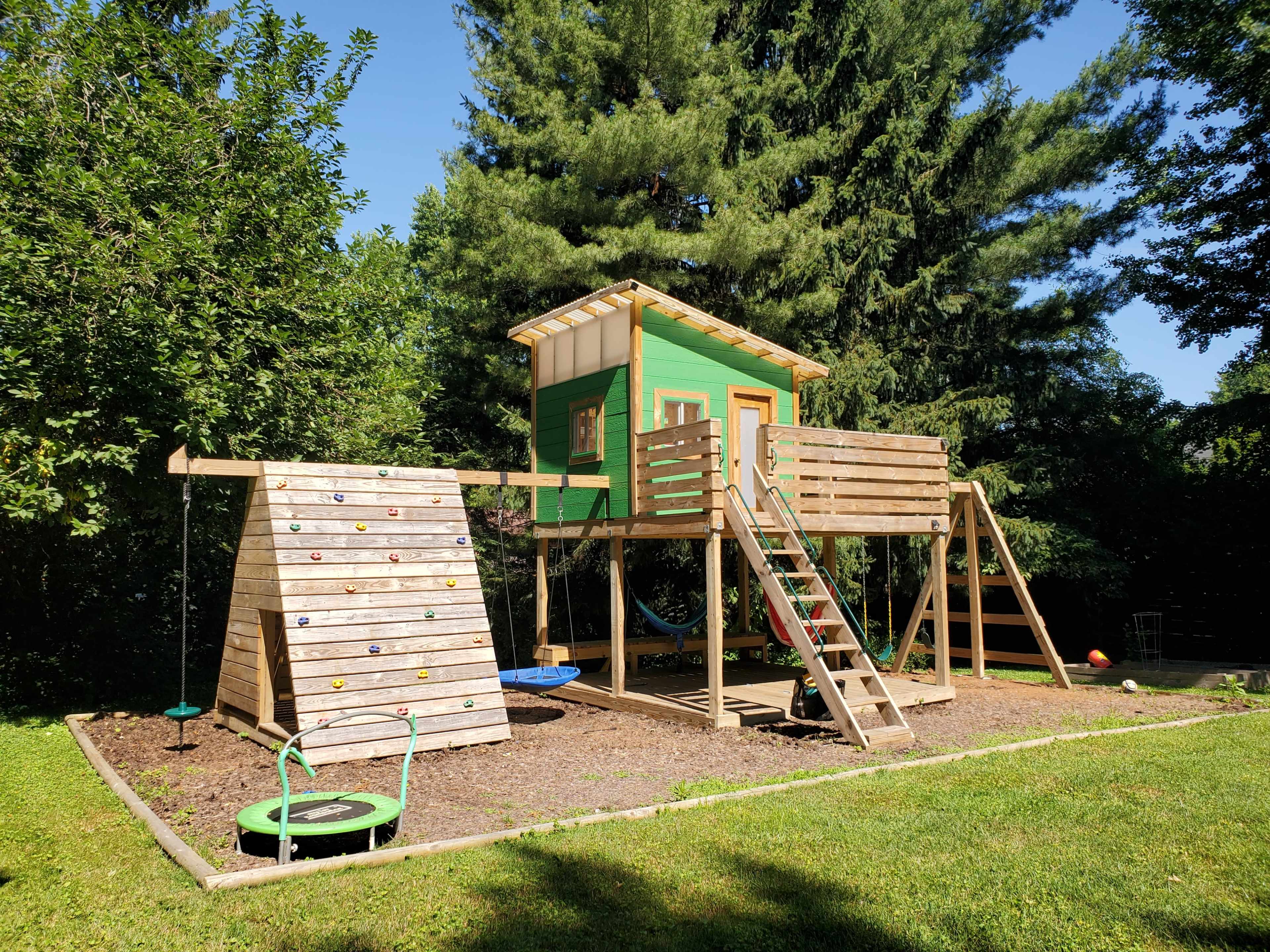 A wooden playset with a green treehouse, a climbing wall, and a swing set, all situated in a grassy yard surrounded by trees.