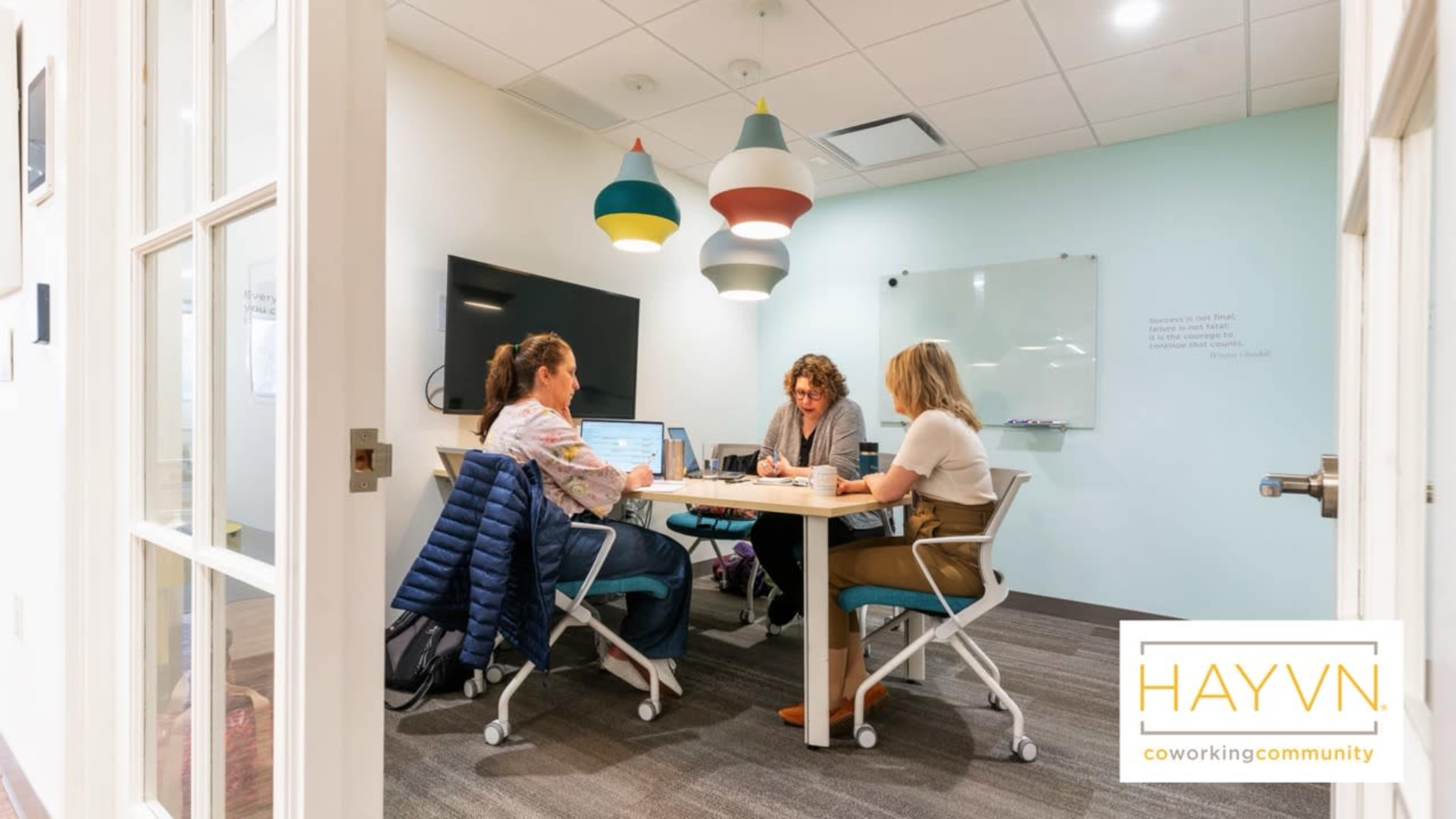 Three women are seated around a table in a brightly lit coworking space, collaborating on laptops with a television screen mounted on the wall in the background.