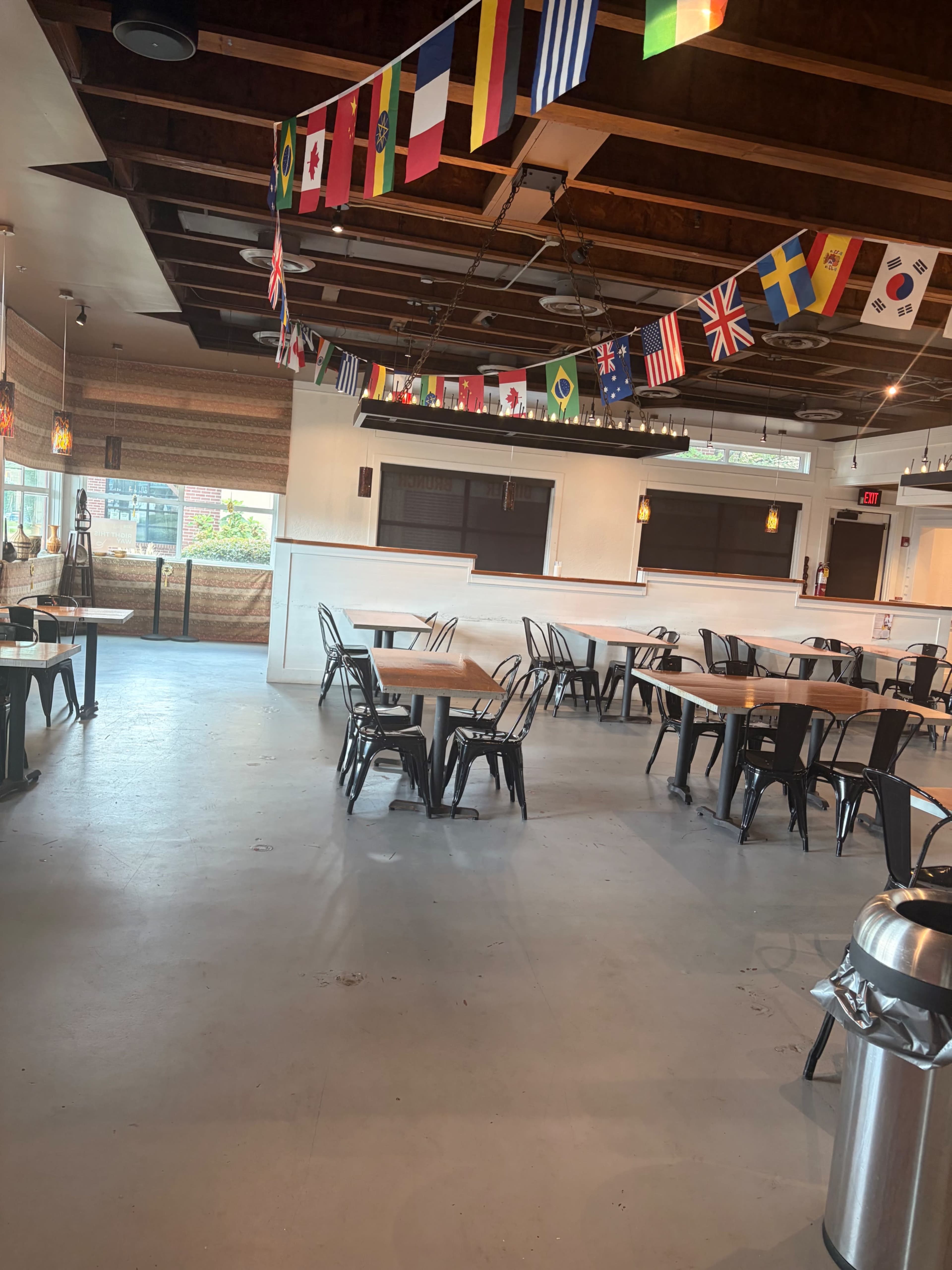 The image shows the interior of a restaurant with empty tables, metal chairs, and a ceiling adorned with various national flags.