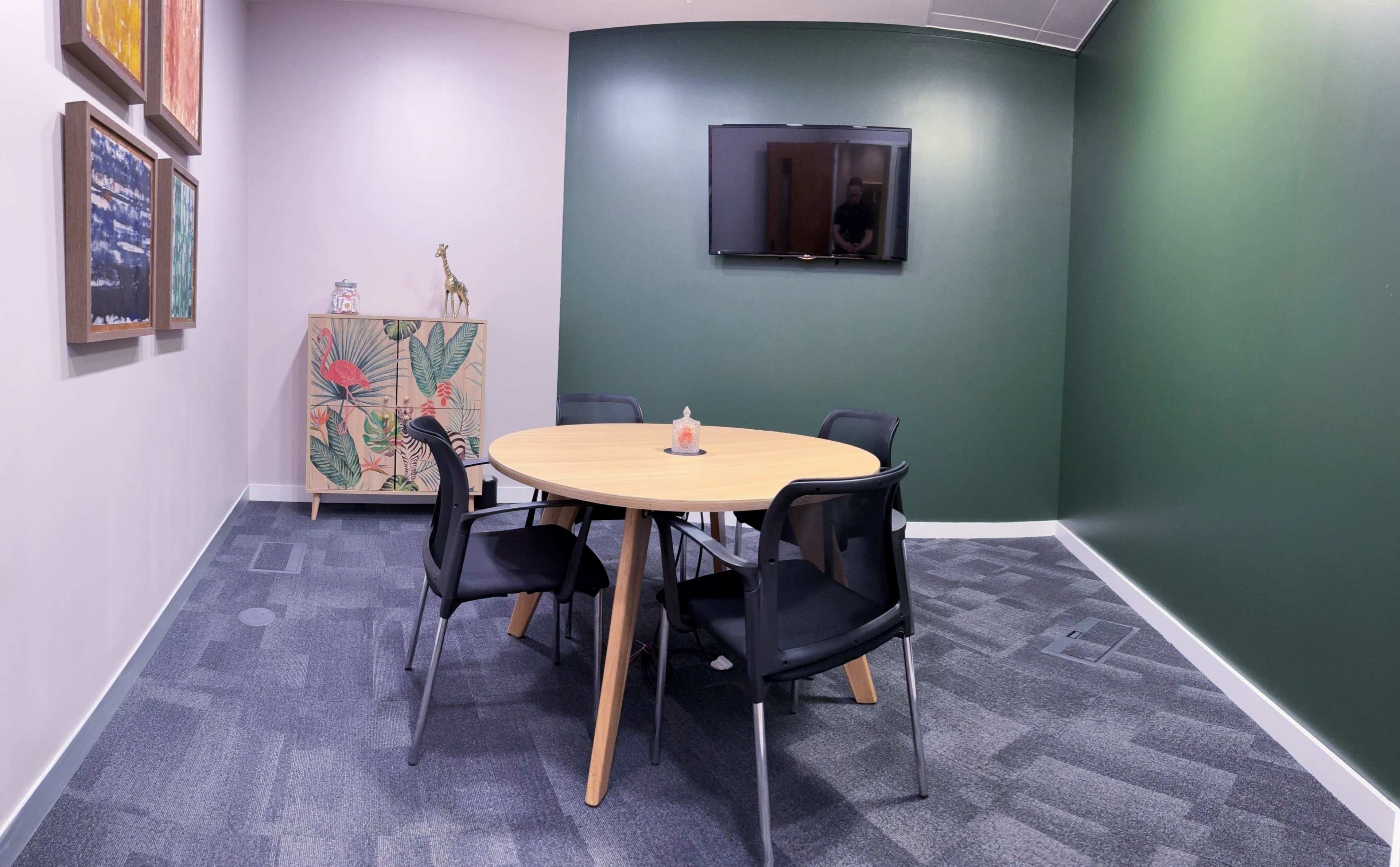 A round wooden table with four black chairs is positioned in a room featuring one green and one light-colored wall, with a television mounted on the wall and a patterned cabinet in the corner.