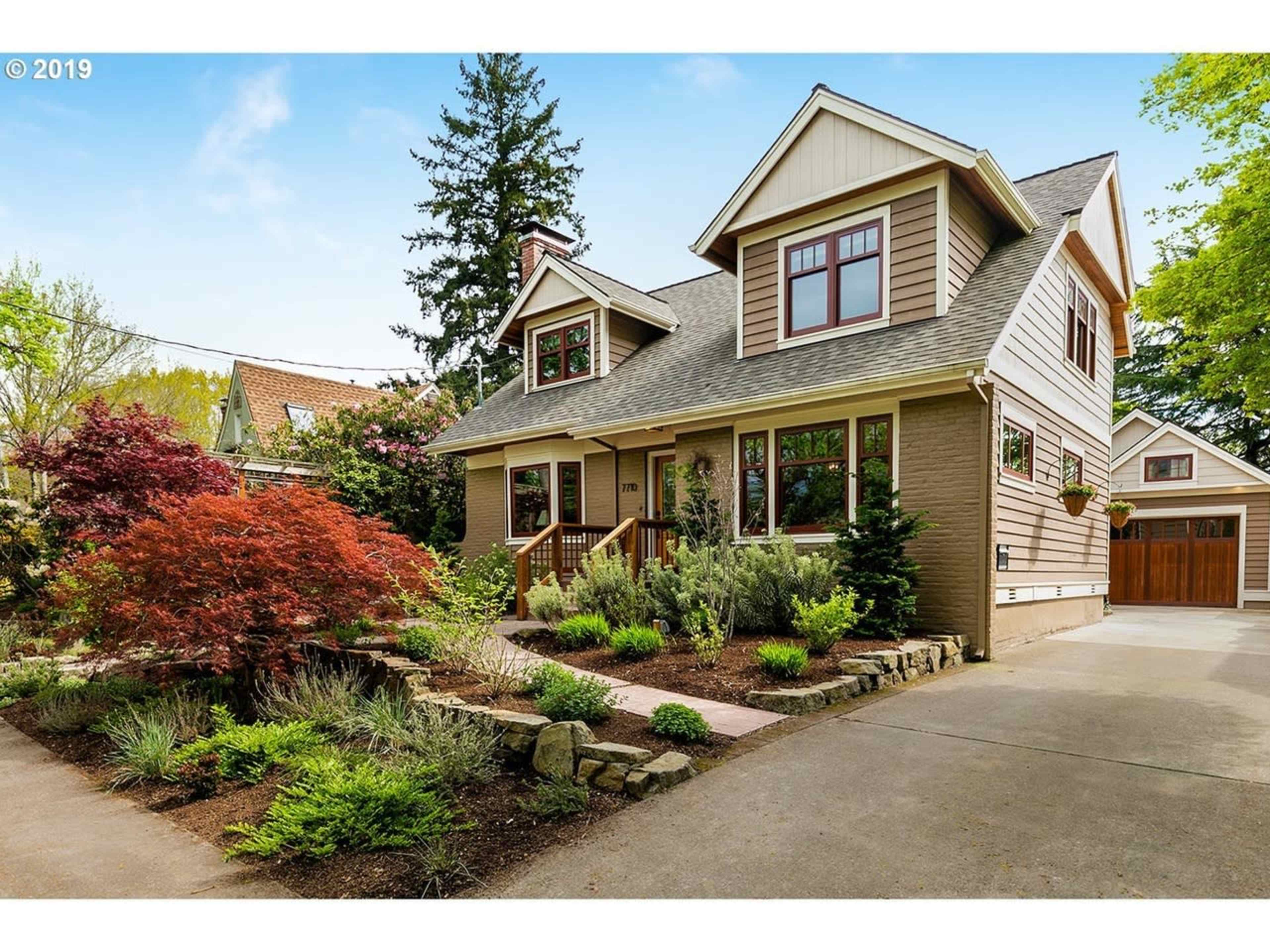 A two-story, craftsman-style house with a landscaped front yard features a mix of shrubs and small trees.