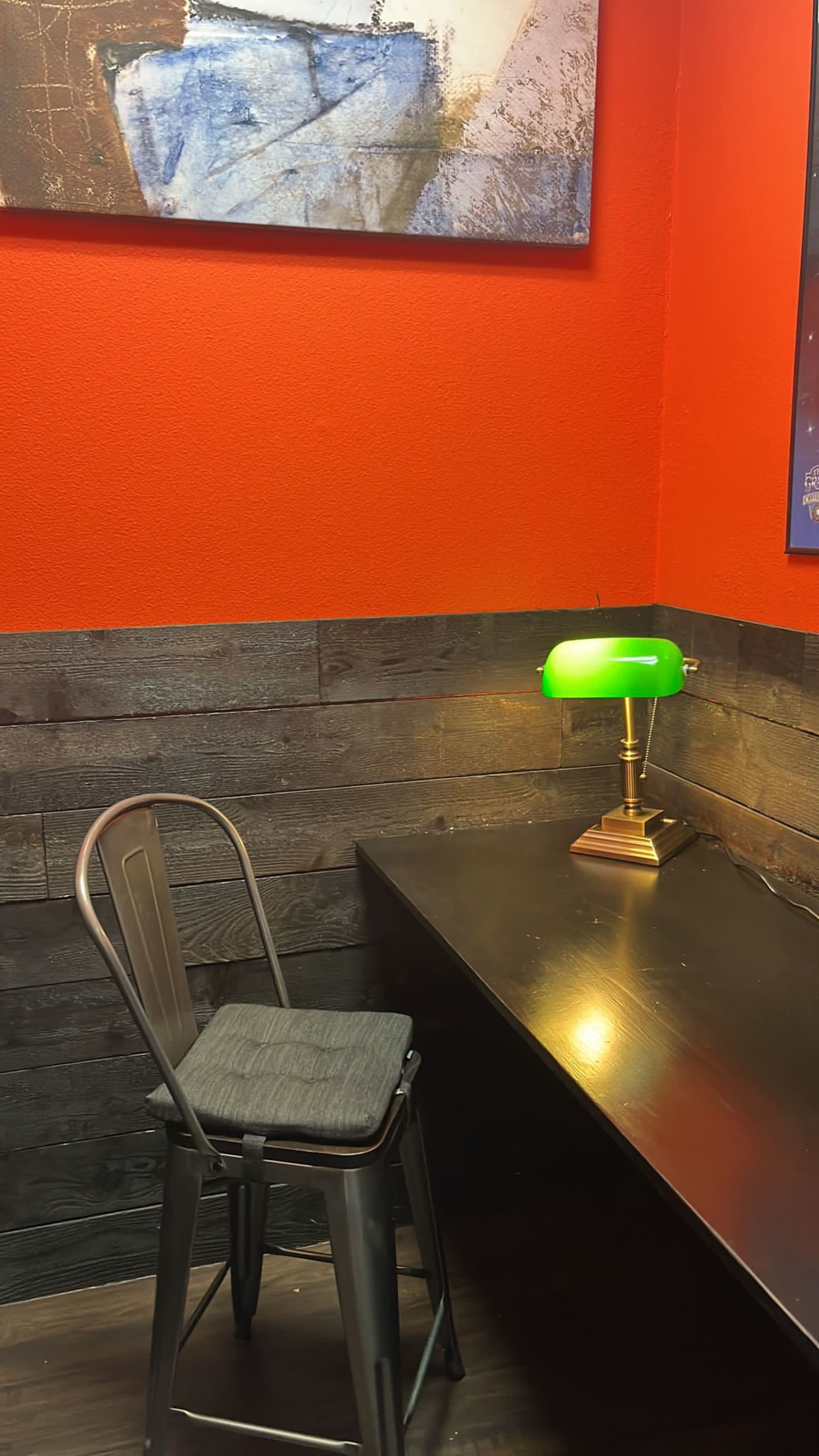 A black metal chair sits beside a dark wooden desk illuminated by a green banker's lamp in a corner with red walls and a framed abstract painting above.