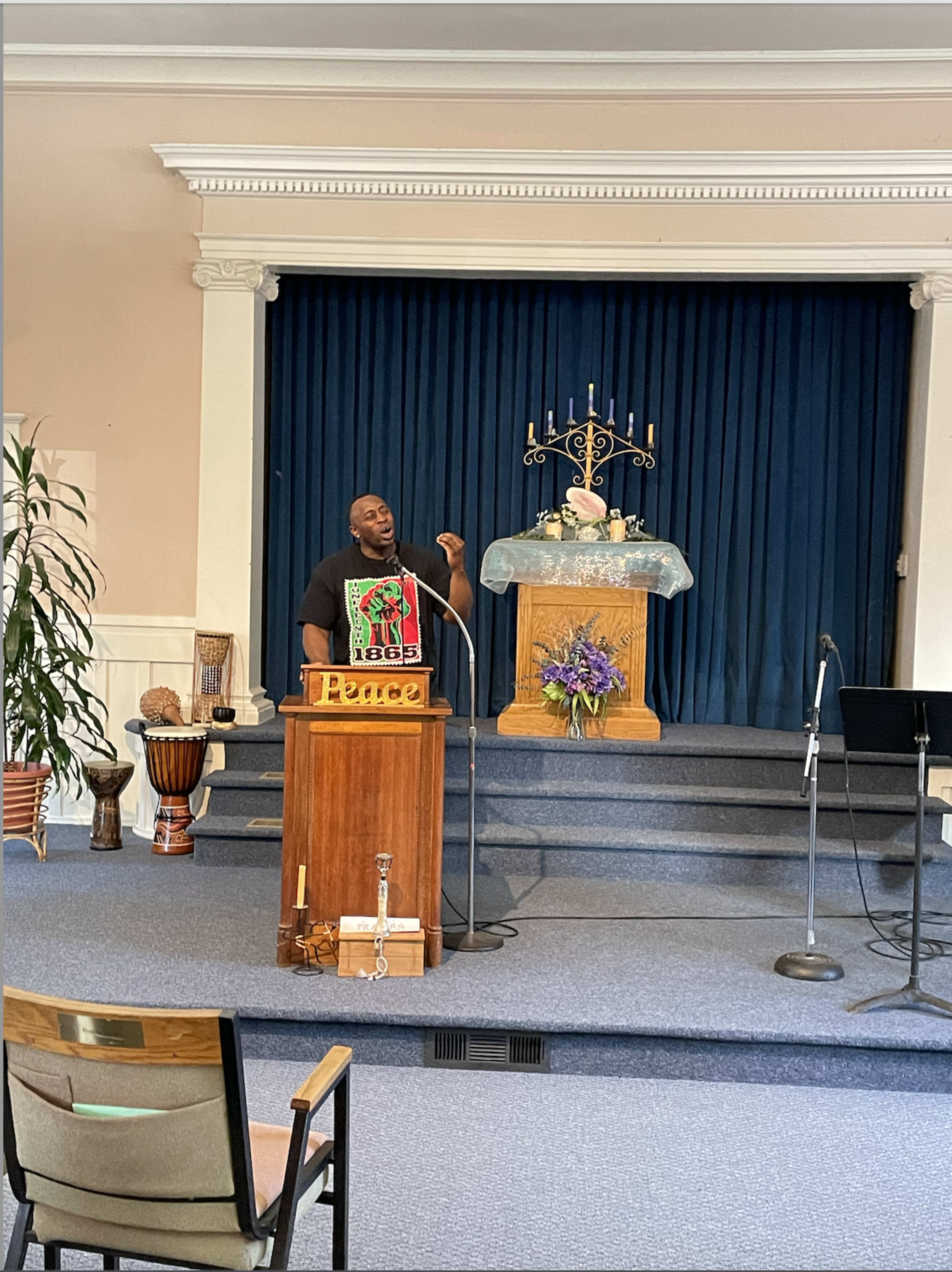 A person stands at a podium labeled "Peace" in a church-like setting with a blue backdrop and decorative altar.