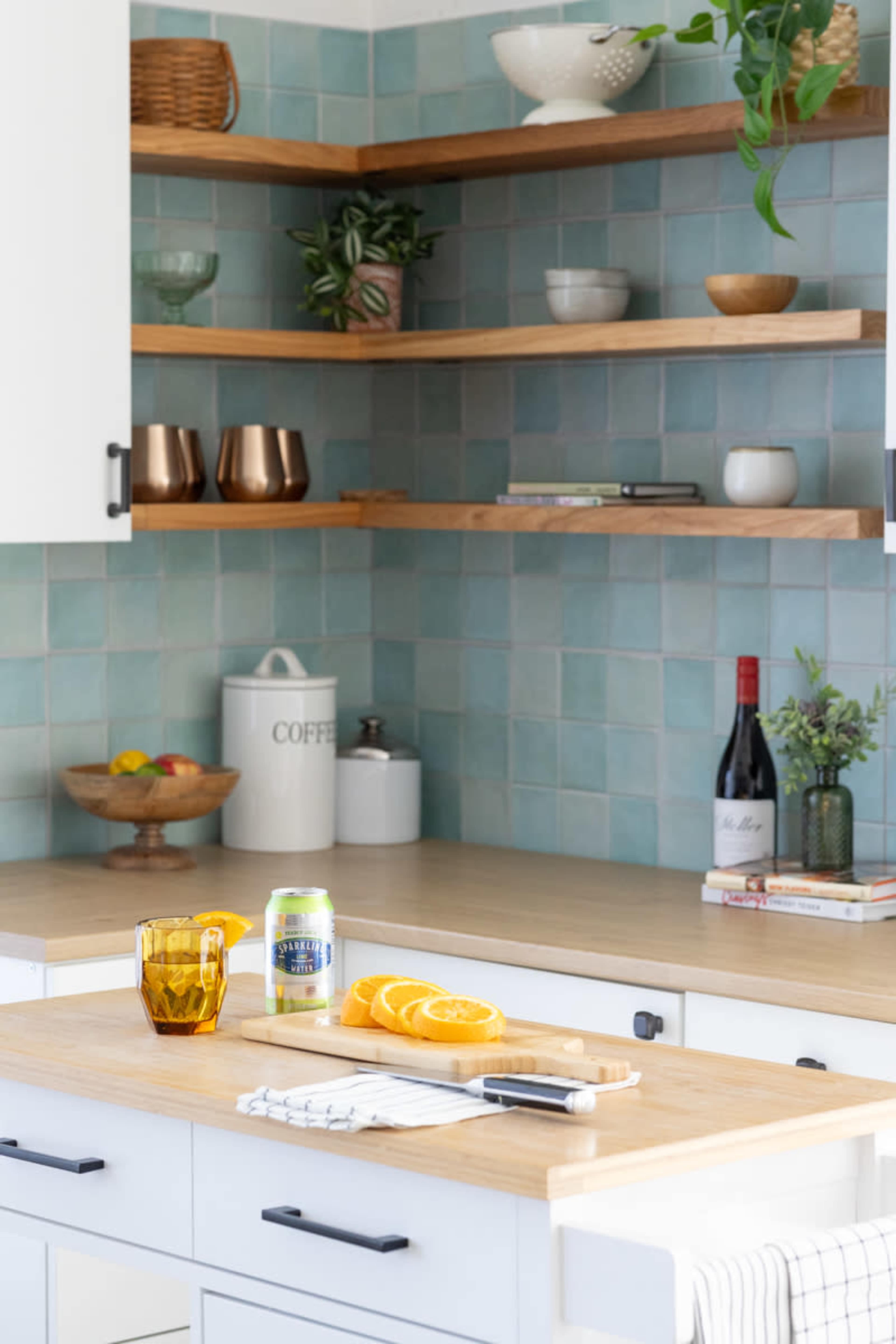 A kitchen countertop features a cutting board with sliced oranges, a knife, and a drink can, set against turquoise tile and wooden shelves displaying various decorative items.