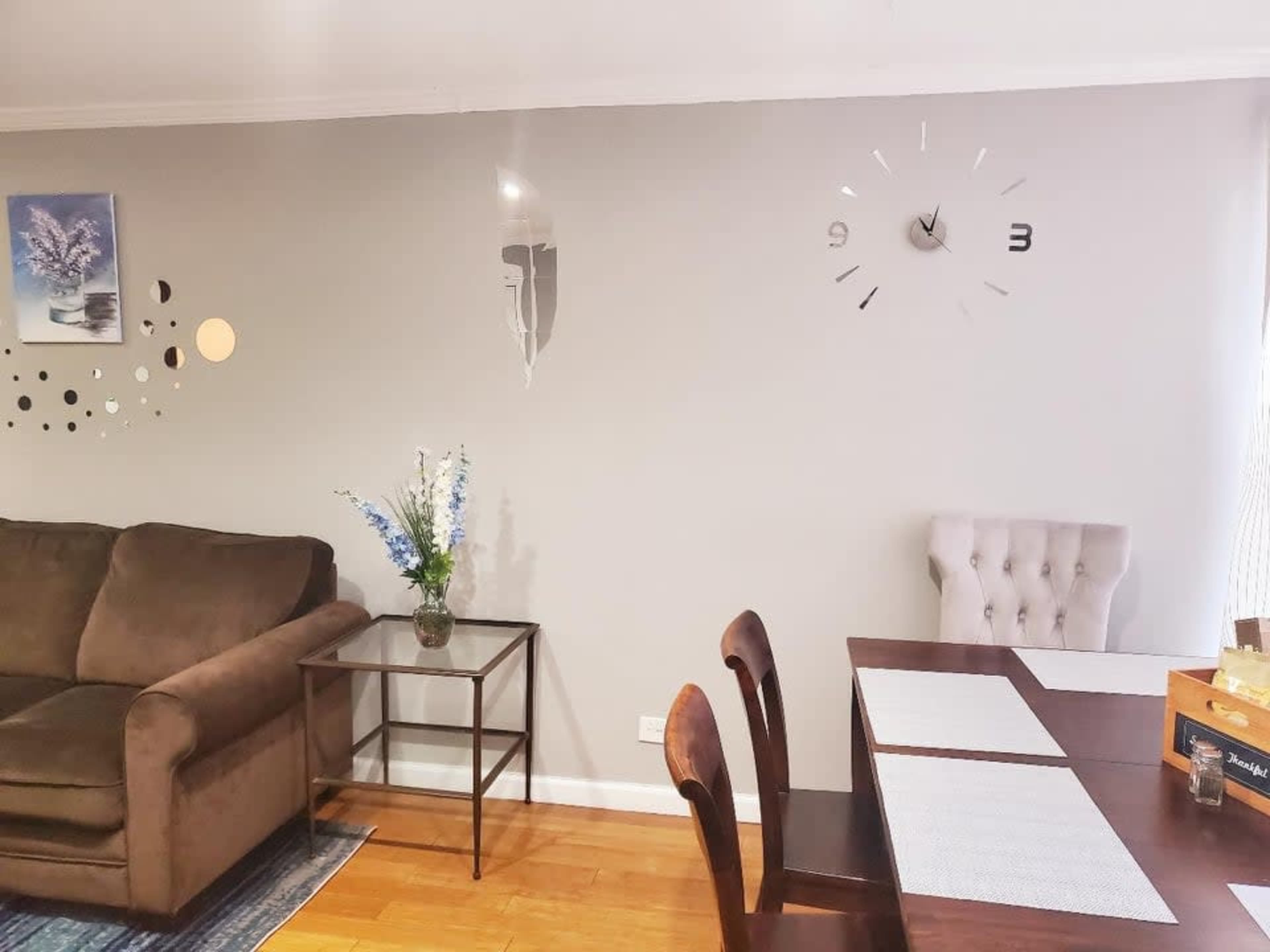 A living space featuring a brown couch, a glass side table, a dining table with two chairs, and a wall clock next to a decorative arrangement of dots and a floral centerpiece.