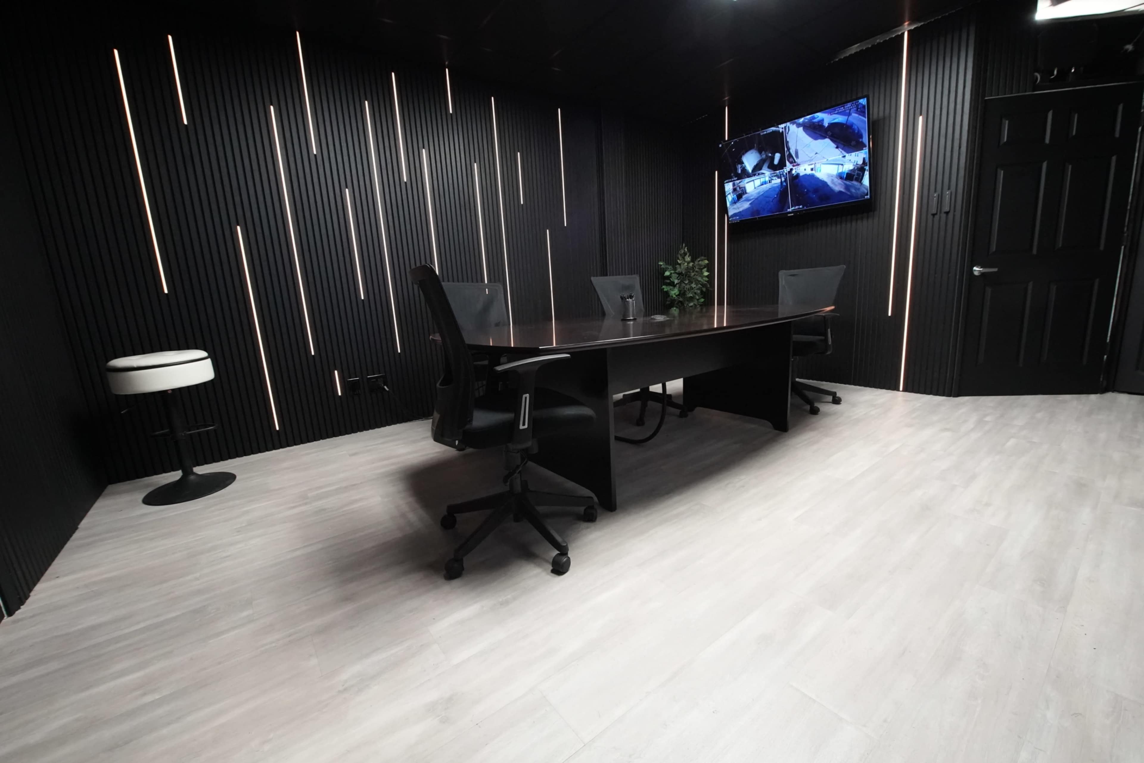 The image shows a modern conference room with a large table, black chairs, and a wall-mounted television, all set against a backdrop of vertical black panels and a light-colored floor.