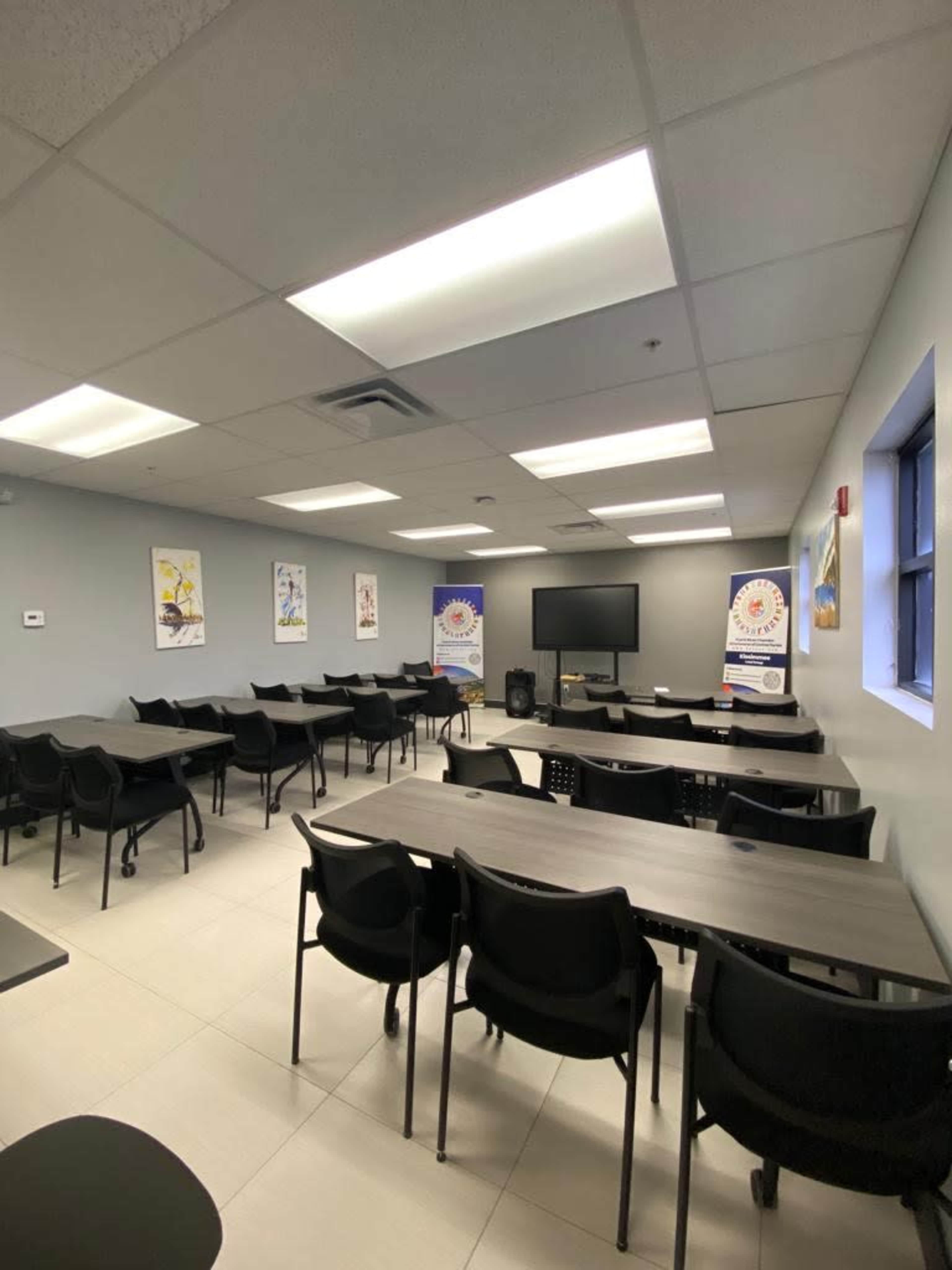 The image shows a classroom or meeting room with multiple rows of black tables and chairs, a large screen at the front, and posters on the walls.