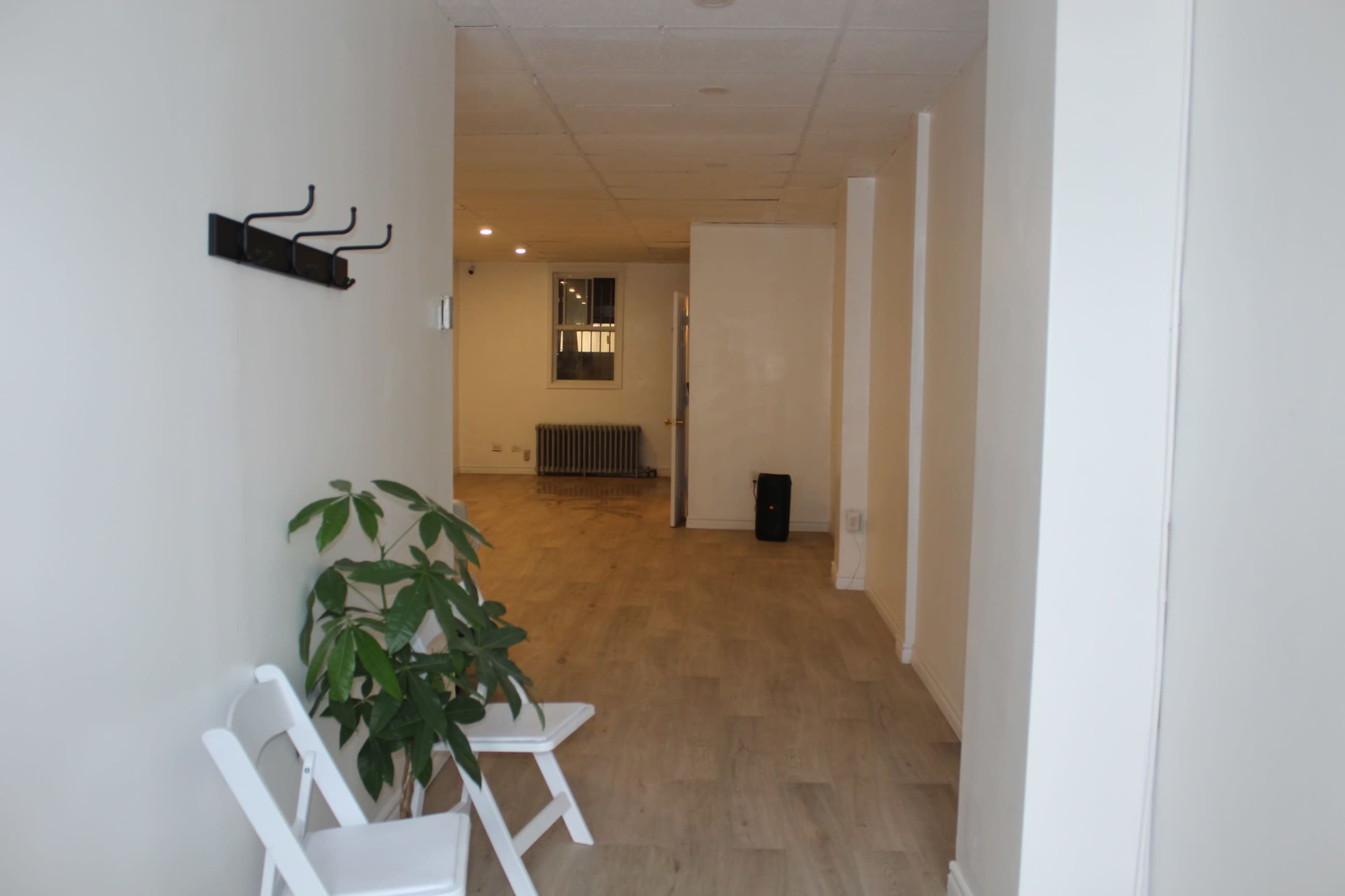 A long, empty hallway with light-colored walls, wooden flooring, a potted plant, and some folding chairs positioned against one side.