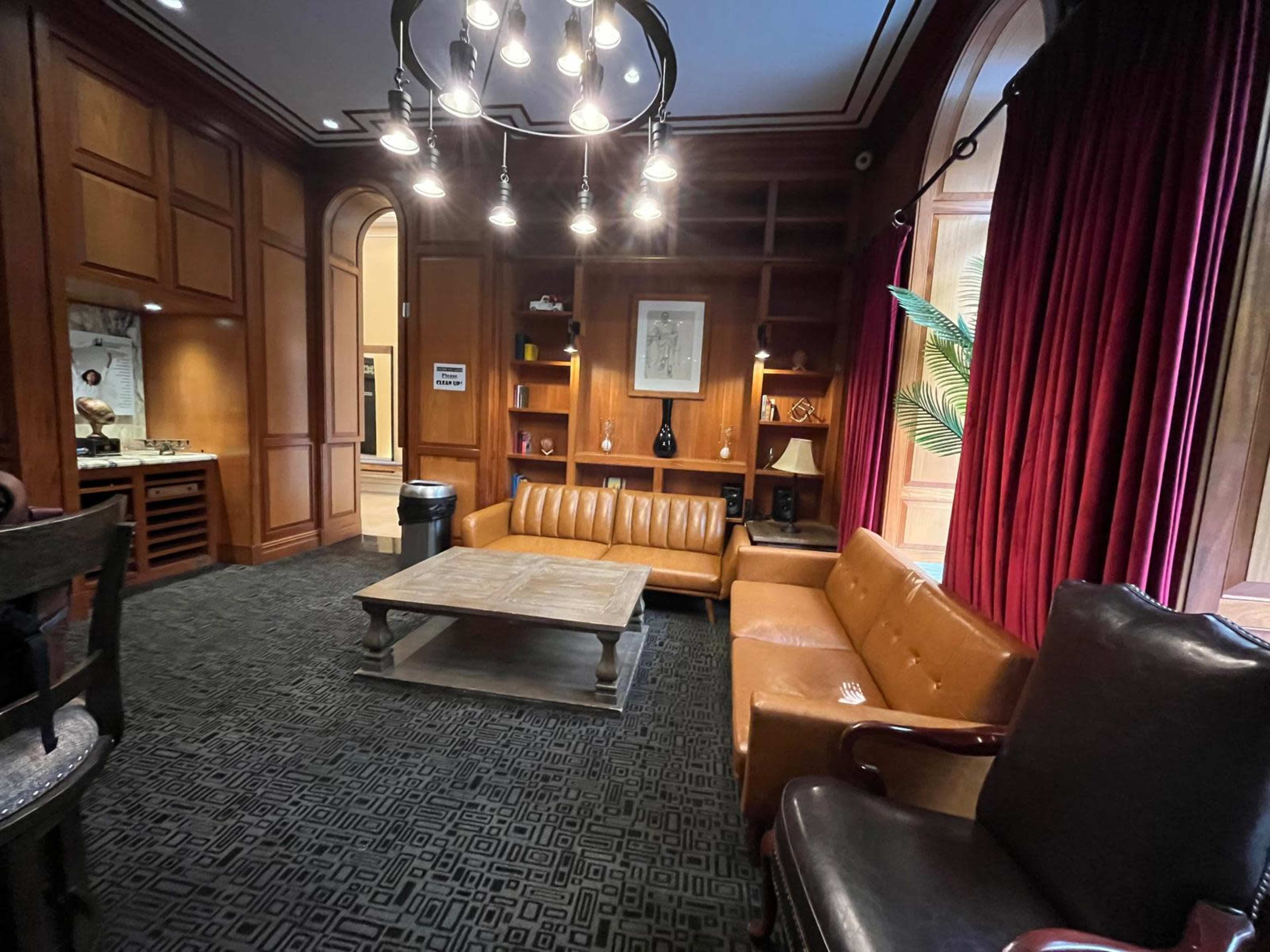 A cozy sitting area with a brown leather couch and armchair, a wooden coffee table, wood-paneled walls, and lush red curtains.