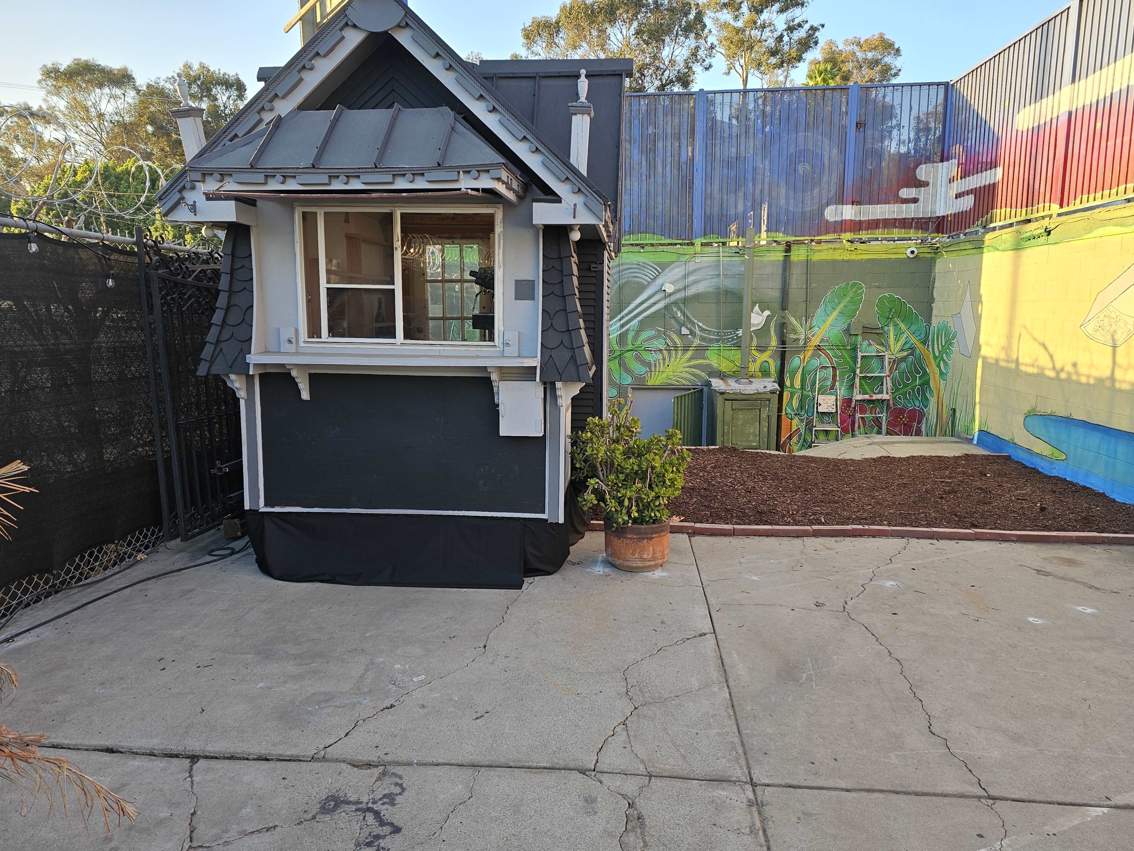 A small black and white house with a sloped roof, situated next to a colorful mural and a gravel area.