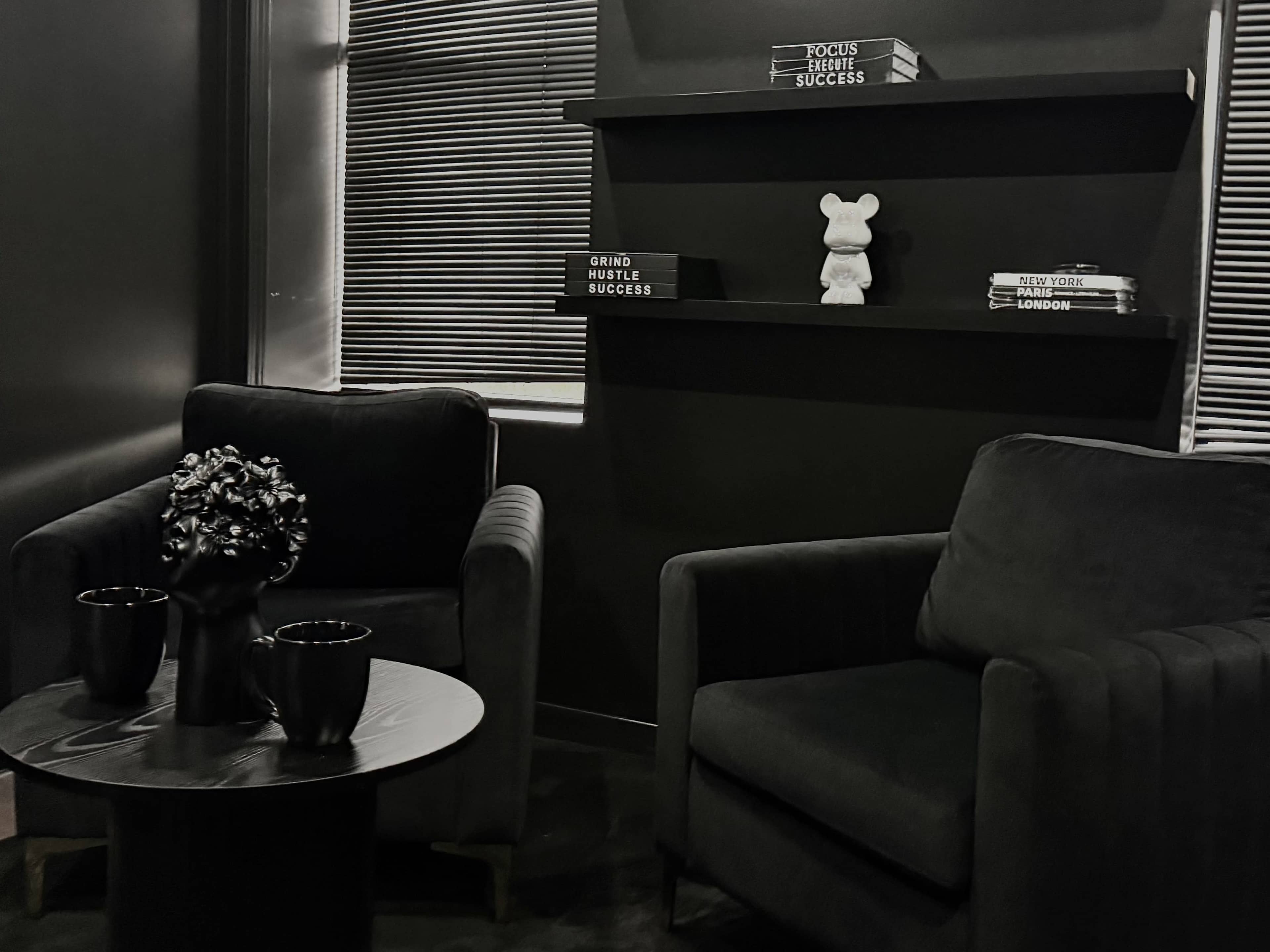 The image shows a contemporary room featuring two dark upholstered chairs, a round coffee table with a vase, and a shelf displaying books and a figurine, all set against a black wall with striped window shades.