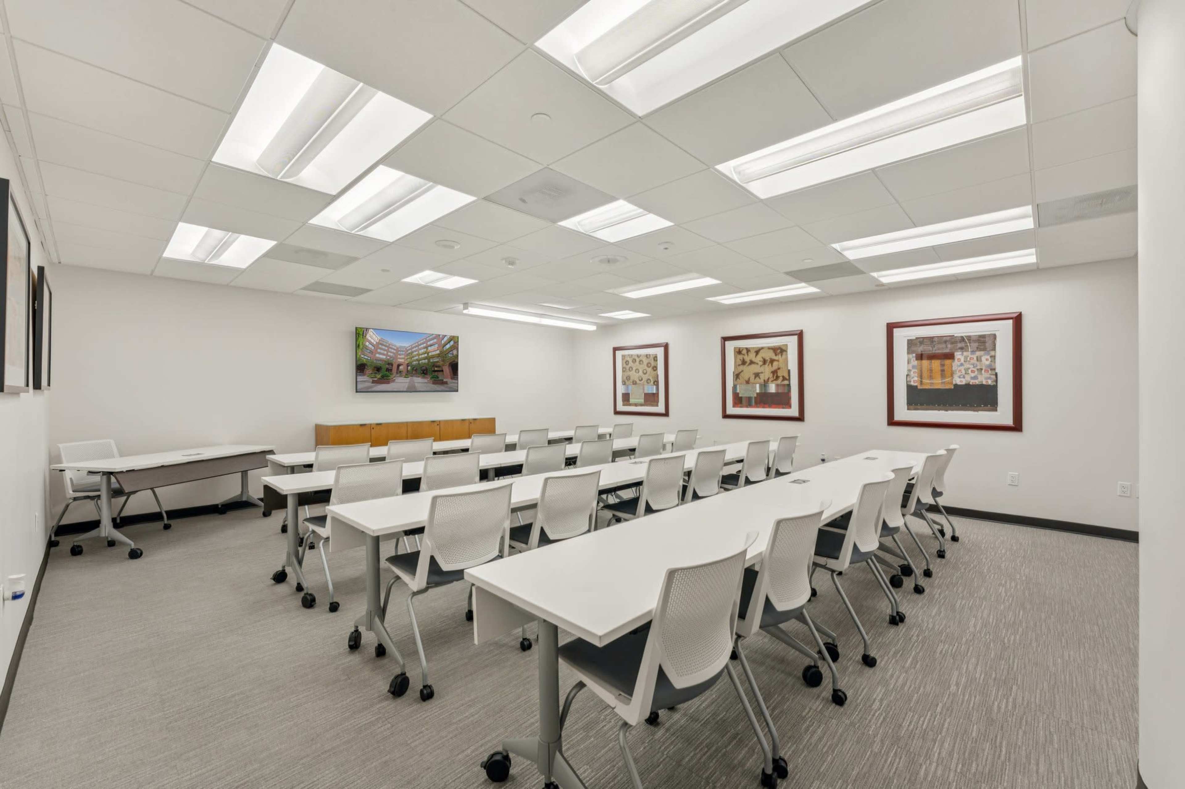 A modern conference room is set up with several white tables and chairs arranged for a meeting, featuring artwork on the walls and a large screen at the front.