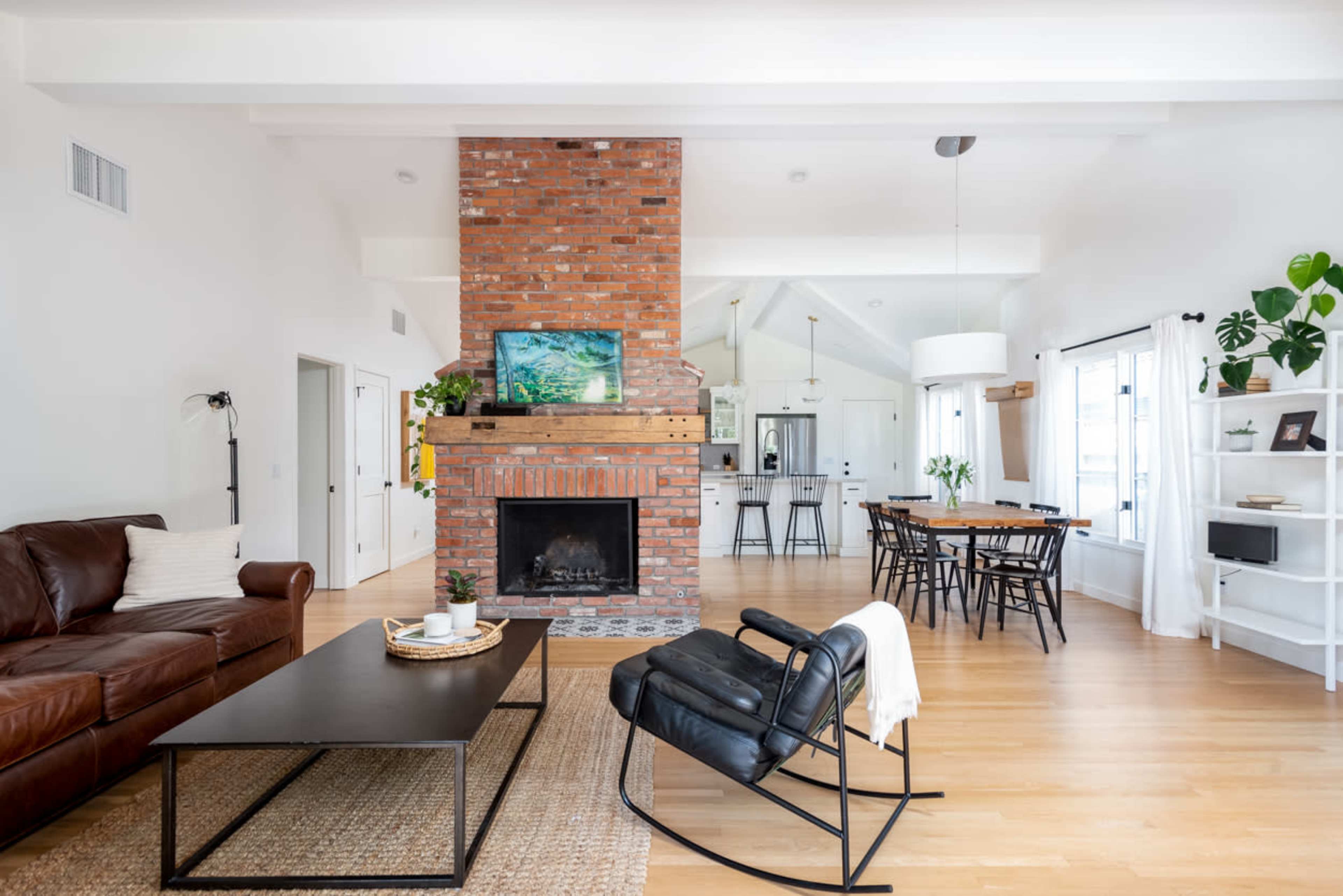 A modern living room with a brick fireplace, a leather sofa, a black coffee table, and a dining area in the background.