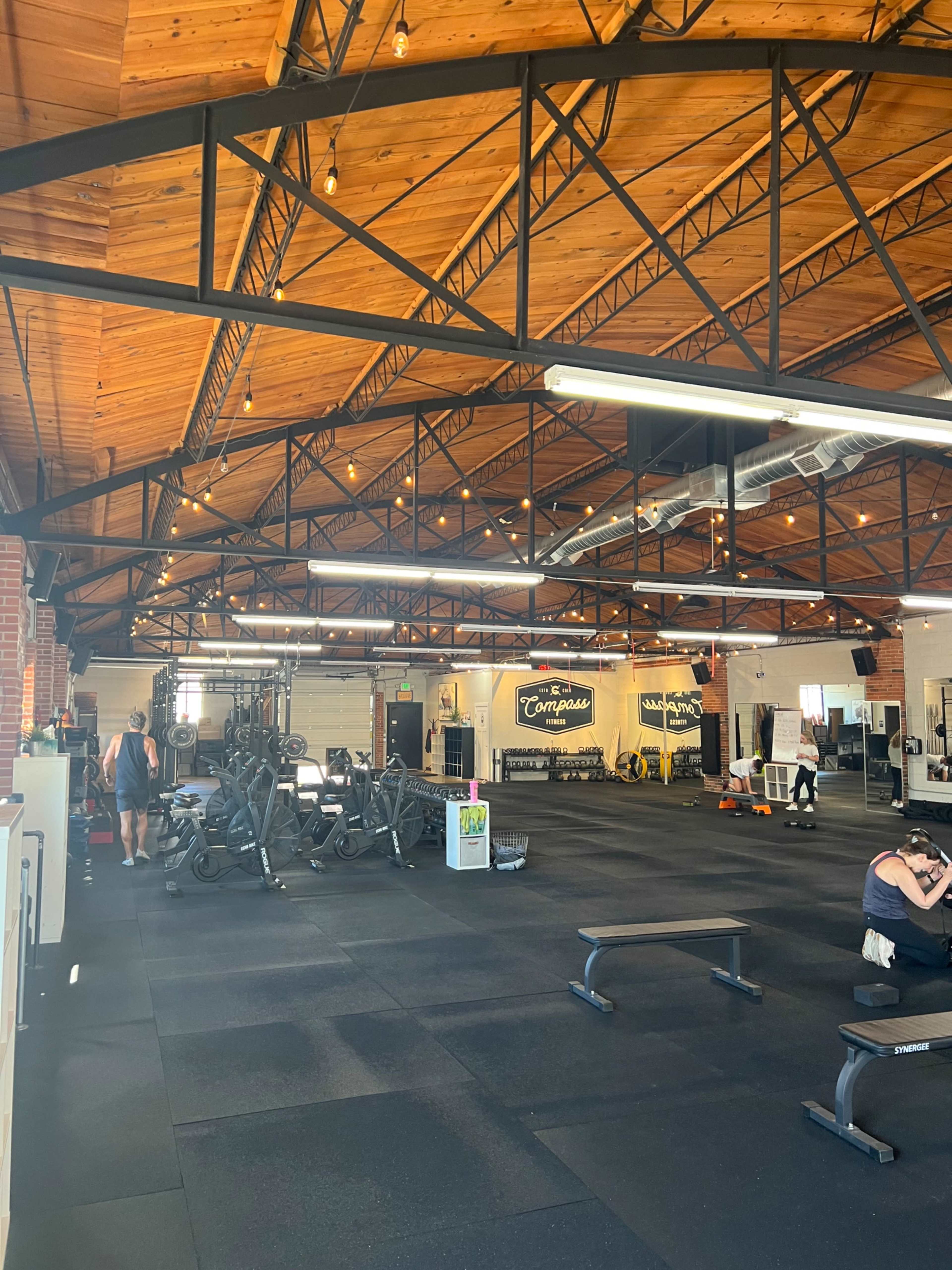 The image shows a spacious gym with high wooden ceilings, various exercise equipment, and individuals engaged in workouts.