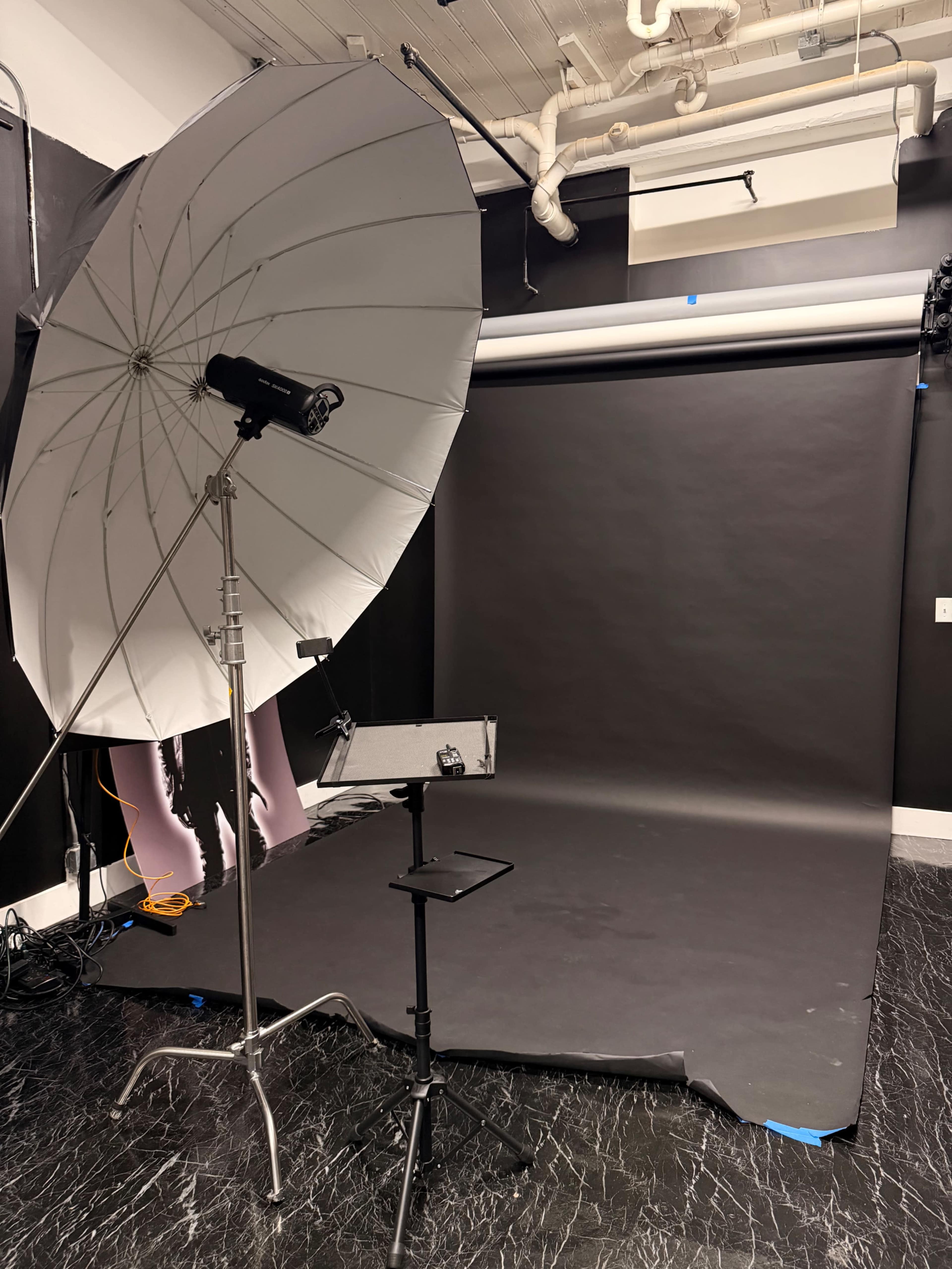 The image shows a photography studio setup with a large white umbrella reflector, a light stand, and a black seamless backdrop on the floor.