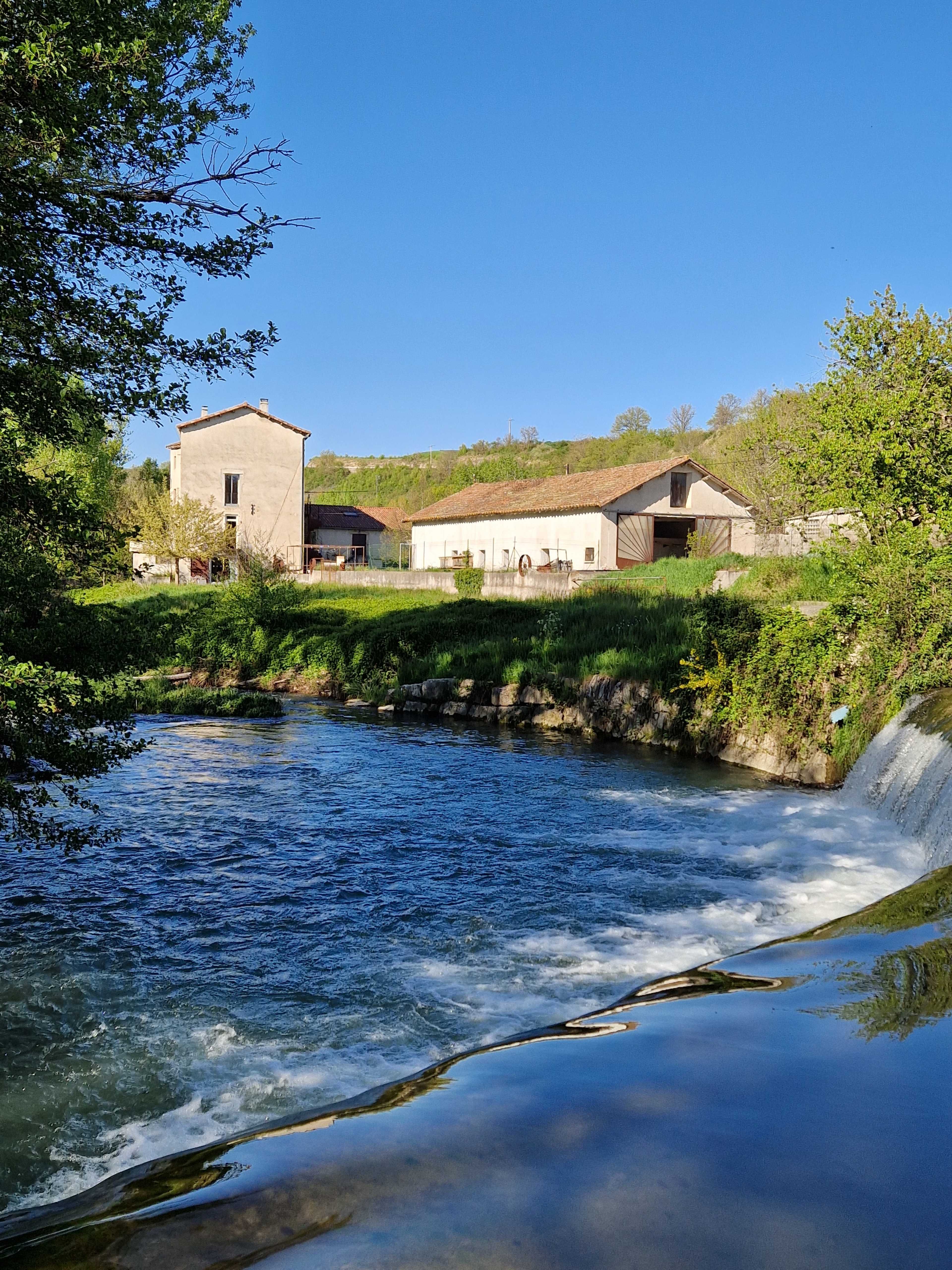 A river flows beside several rustic buildings set against a hillside under a clear blue sky.
