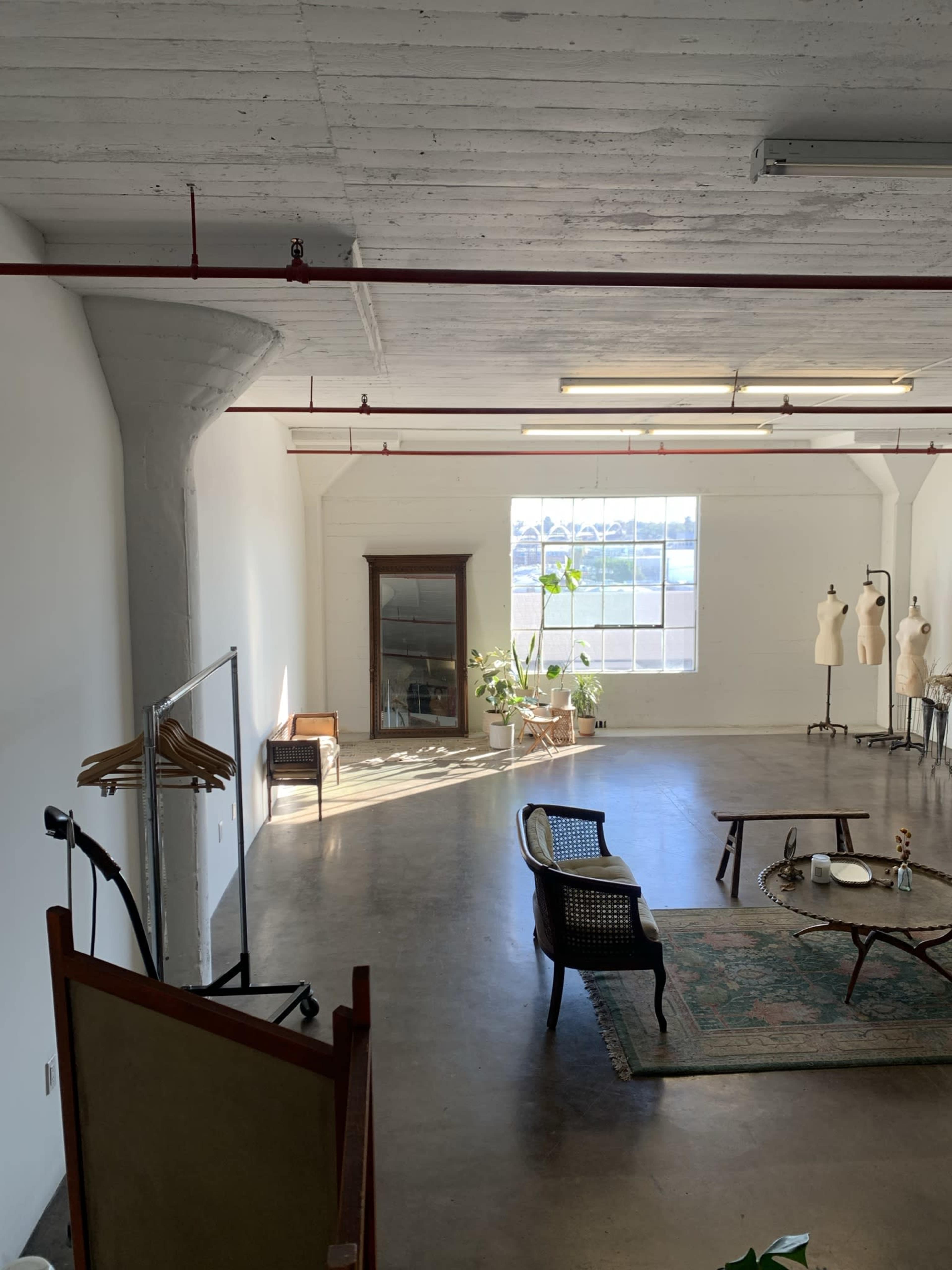 A spacious, well-lit studio interior with a large window, several plants, vintage furniture, and a clothing rack.