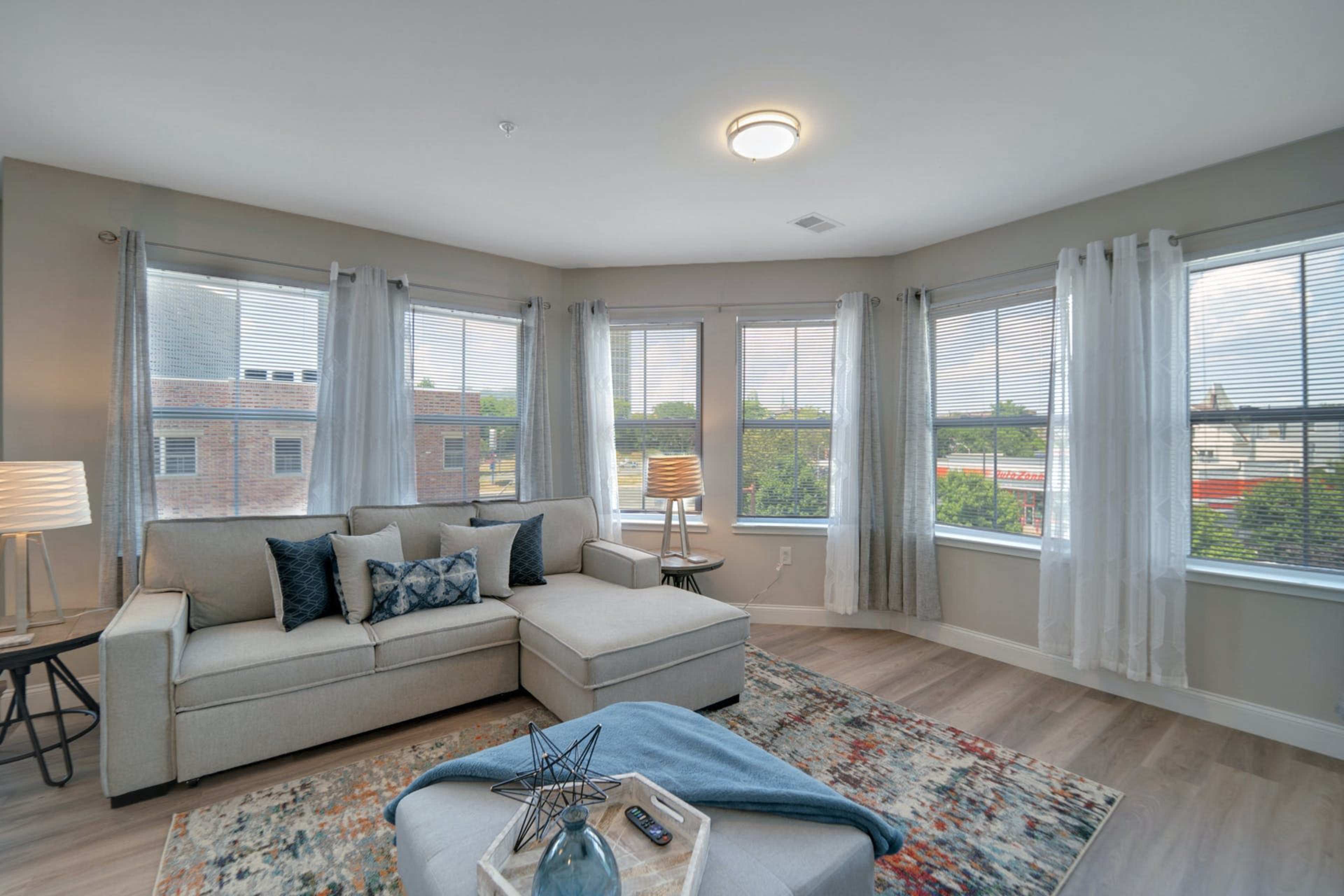 A modern living room features a sectional couch, a coffee table, and large windows with sheer curtains allowing natural light to enter.
